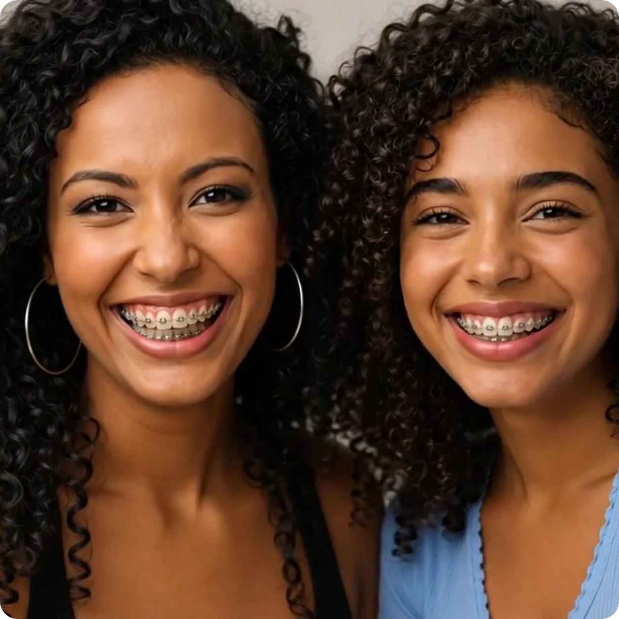 Two young women with curly hair smiling, showing braces on their teeth, wearing casual tops.