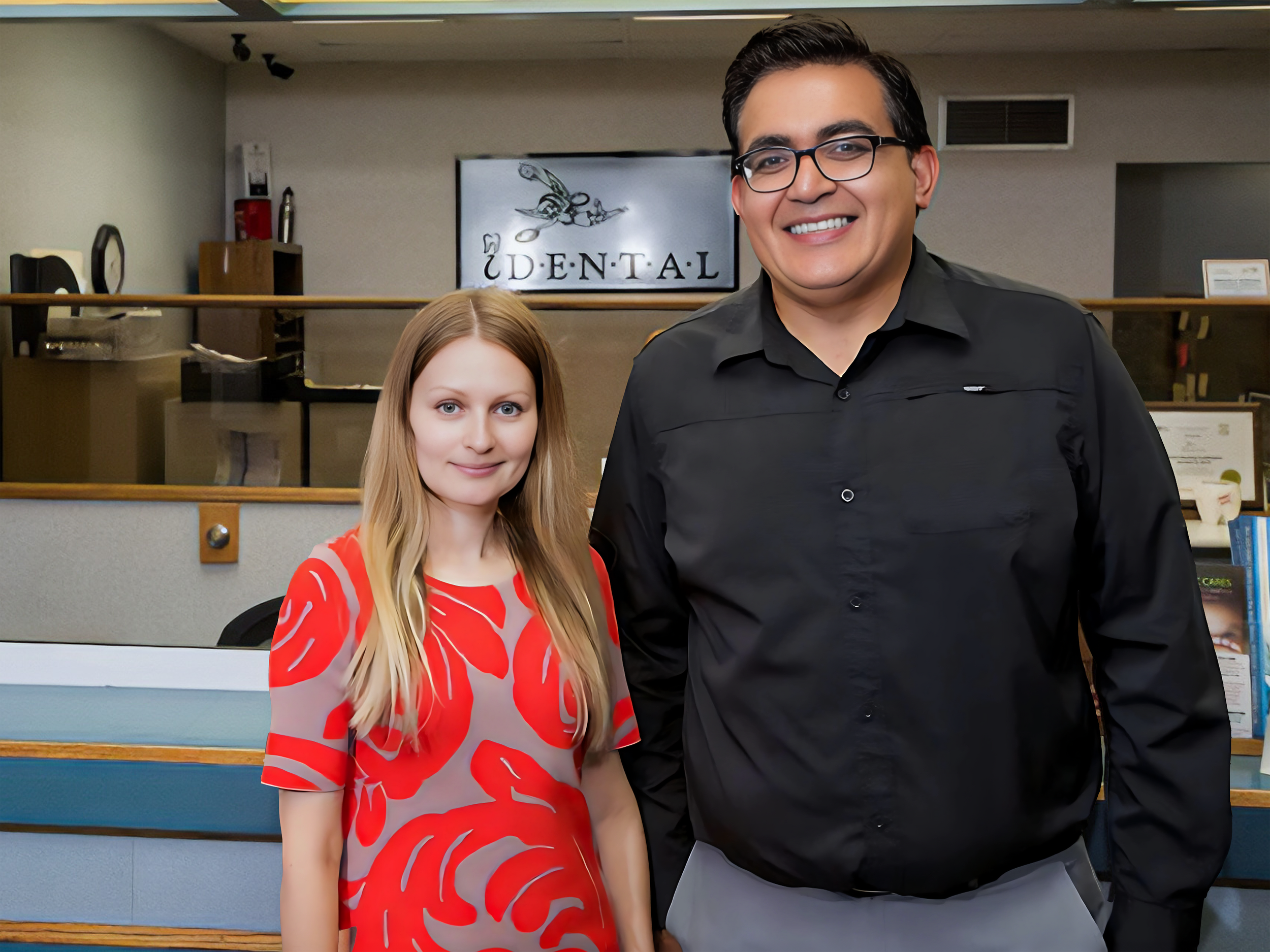 A woman with long blonde hair wearing a red and beige patterned dress and a man with dark hair, glasses, and a black button-up shirt standing at a dental office reception area.