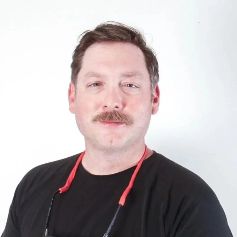 A man with light skin, brown hair, and a mustache, dressed in a black shirt, standing against a plain light background.
