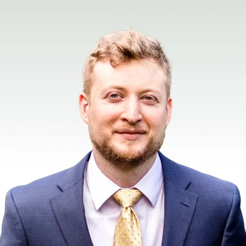 Portrait of a young man with blond hair and a beard, wearing a navy blue suit, white shirt, and a gold tie, smiling slightly against a light background.