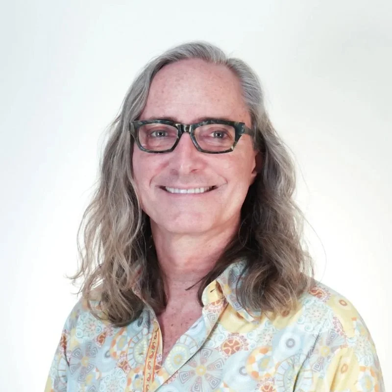 Portrait of a man with long gray hair, glasses, smiling, wearing a colorful, patterned shirt, against a plain light background.