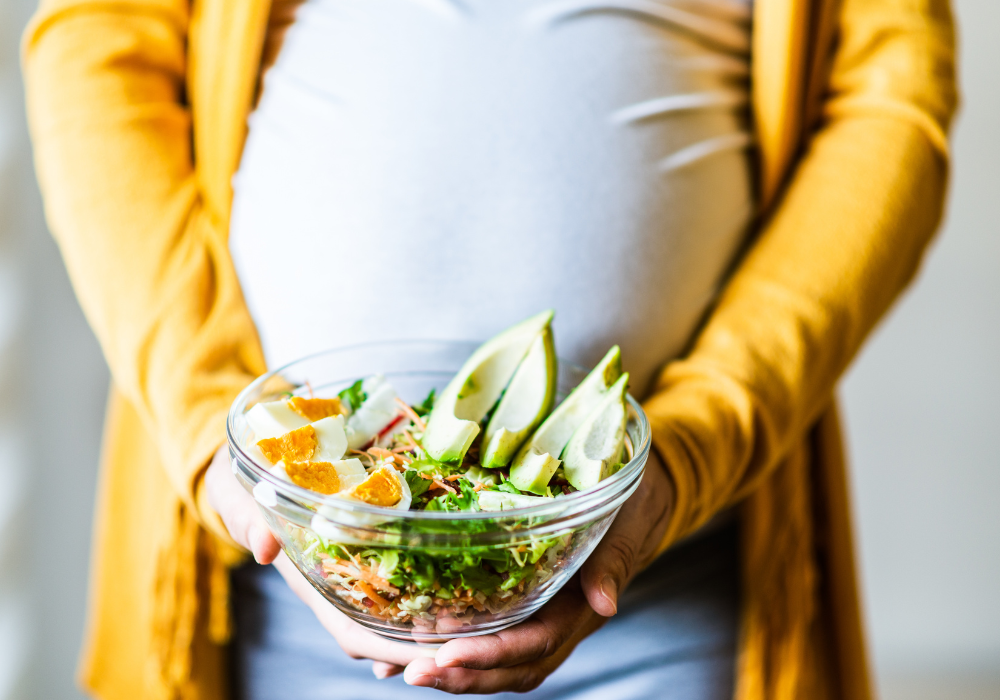 This image shows a pregnant woman in a yellow sweater holding a bowl of real foods that promote optimal nutrition during pregnancy.