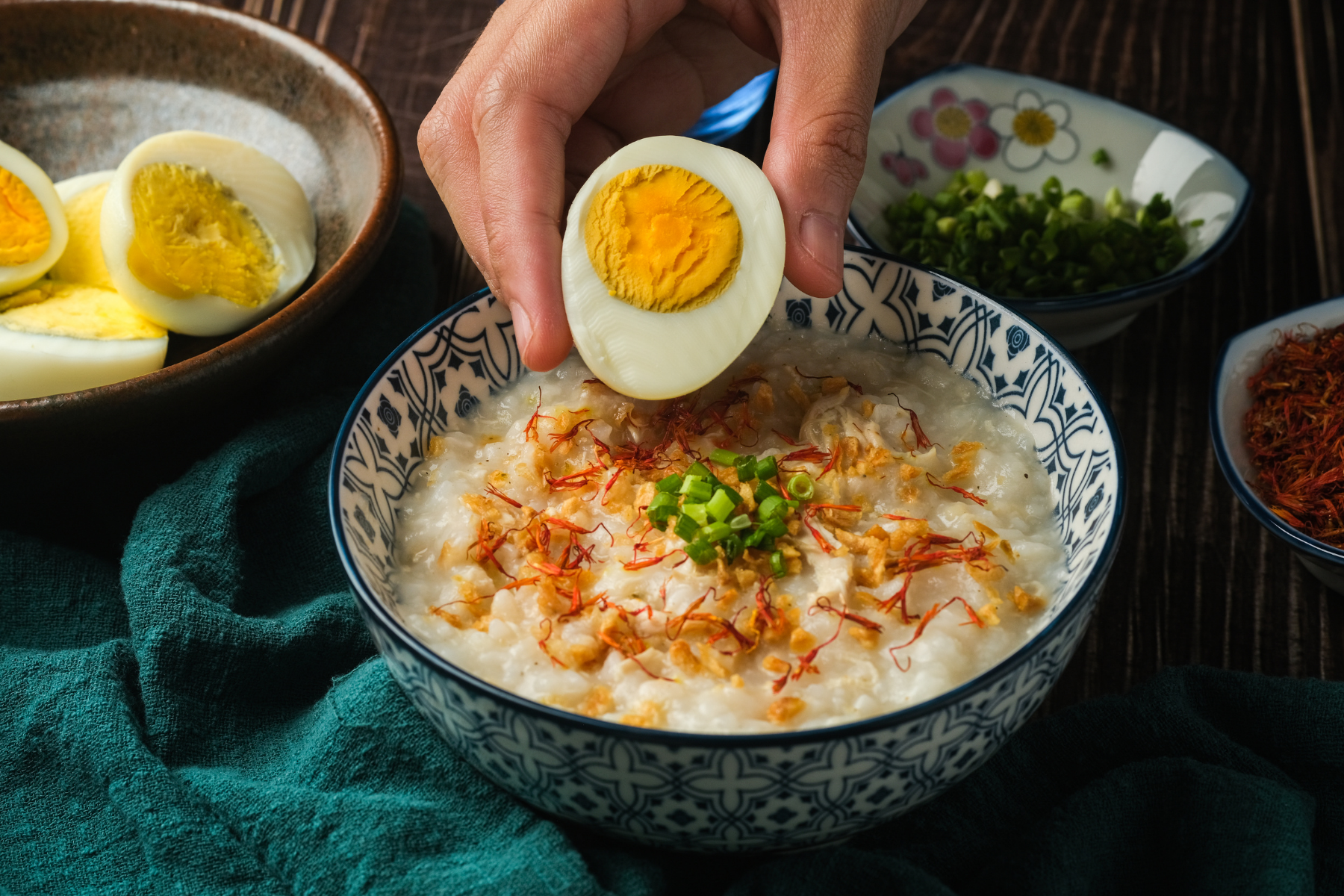 Person adding a halved boiled egg to a bowl of congee with green onions and saffron strands, with side bowls of chopped green onions, shredded meat, and additional boiled eggs on a dark wooden surface.