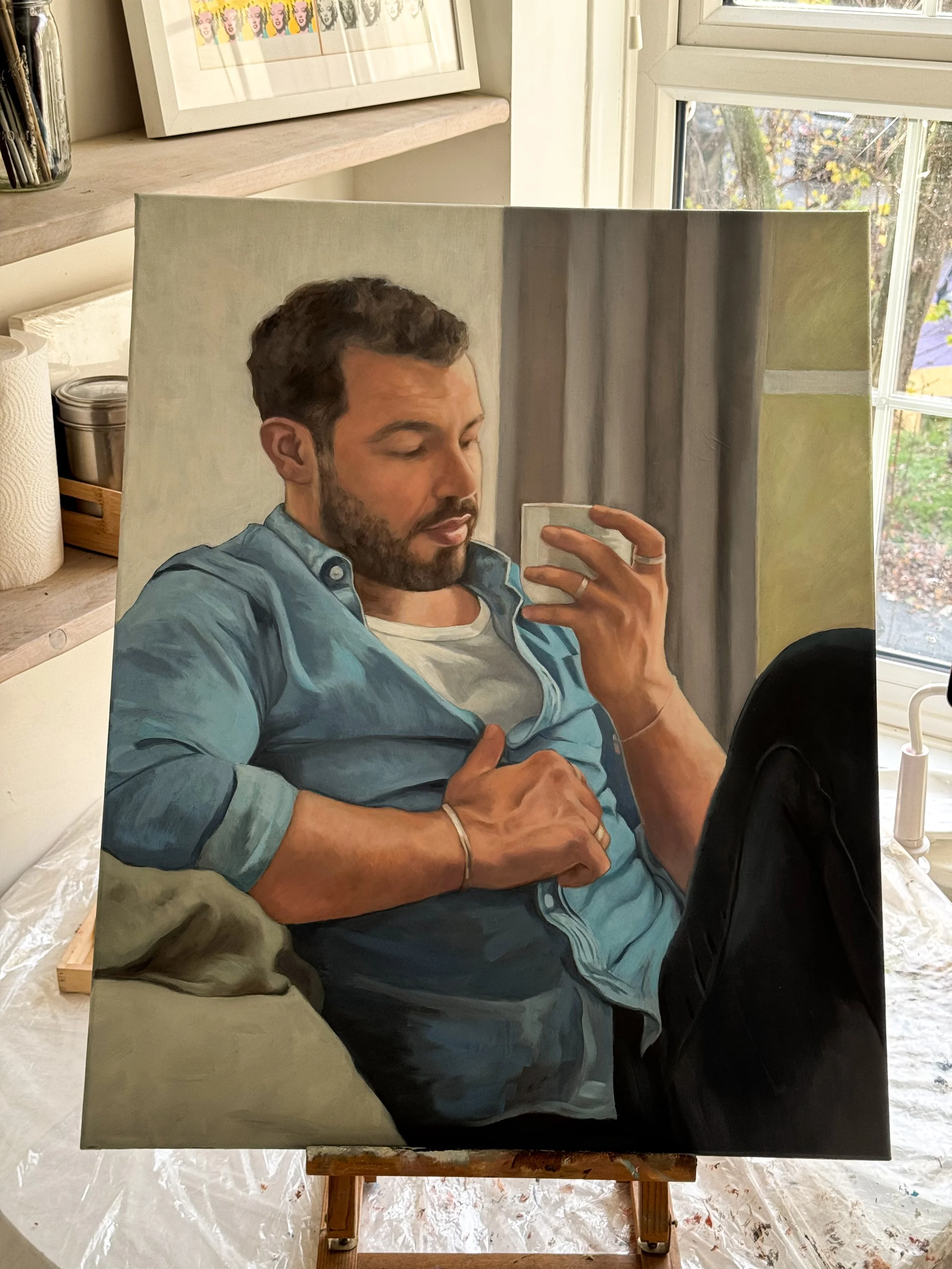 A realistic painted portrait of a man with dark hair and a beard, wearing a blue denim shirt over a white t-shirt, sitting indoors and taking a selfie with a smartphone.