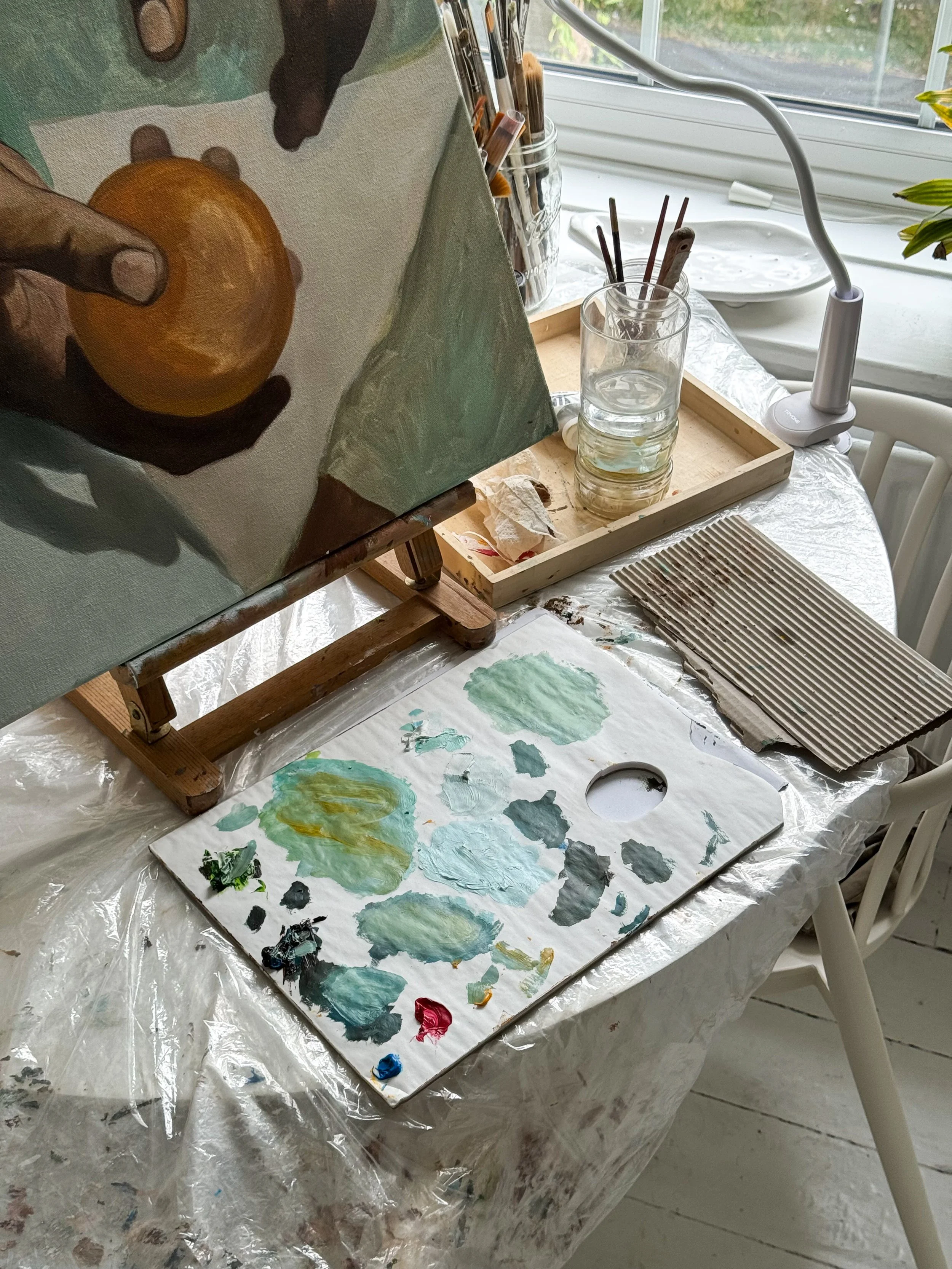 Art workspace with a small canvas on an easel, a paint palette with green, blue, and yellow paint, paintbrushes in a glass, and a water jar, on a table covered with plastic, near a window.