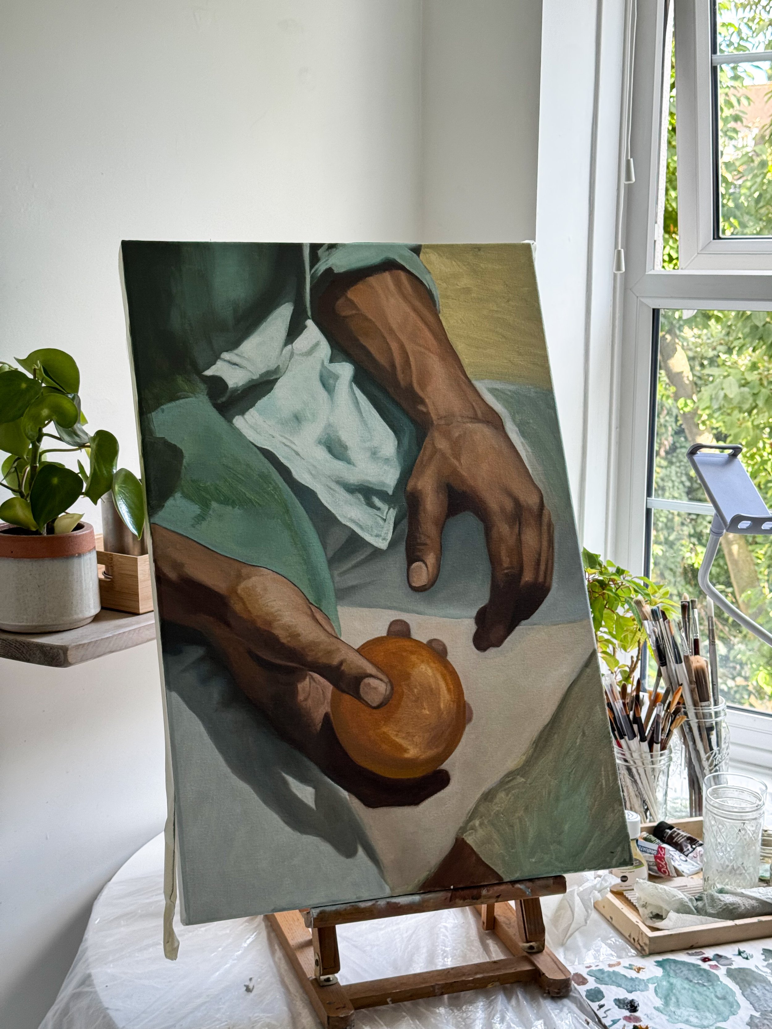 A painting of hands holding a small orange object on a table, set up on an easel in a bright room with a window and artist supplies nearby.