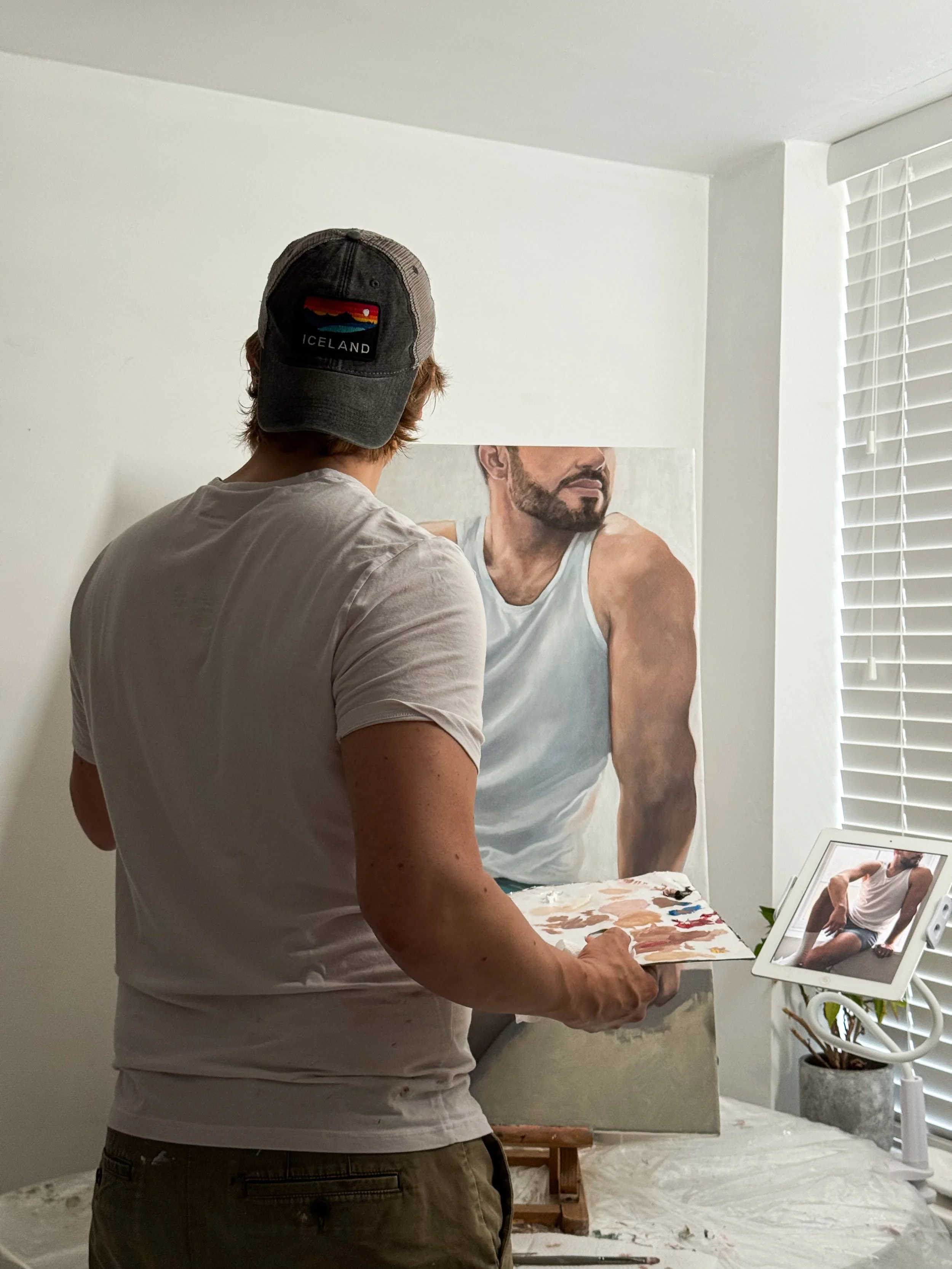 An artist standing in front of a painting of a man wearing a white tank top, while looking at their reflection in a tablet on a stand.