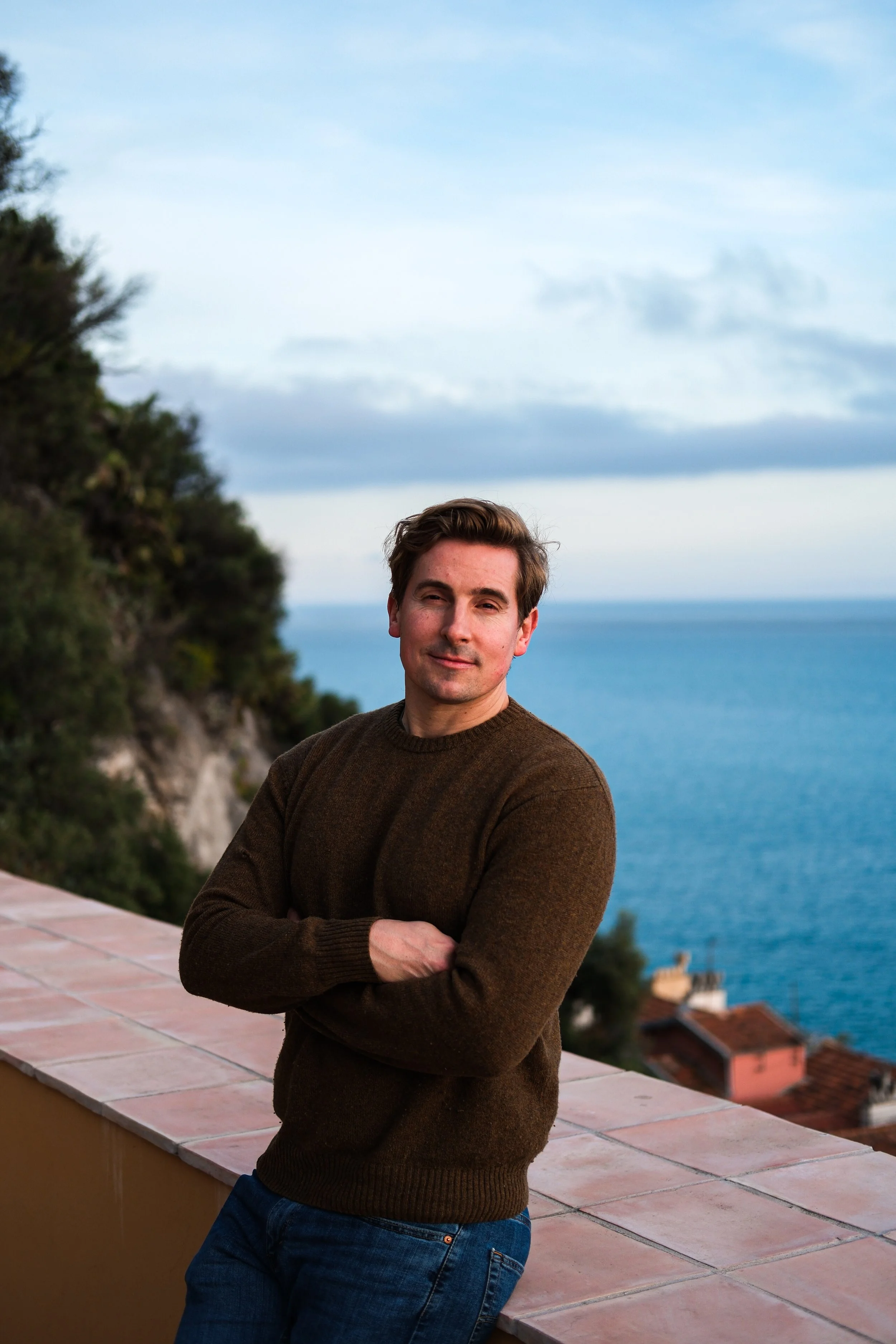 A man with brown hair, wearing a brown sweater and blue jeans, standing with arms crossed on a balcony with a view of the sea and coastal houses in the background.