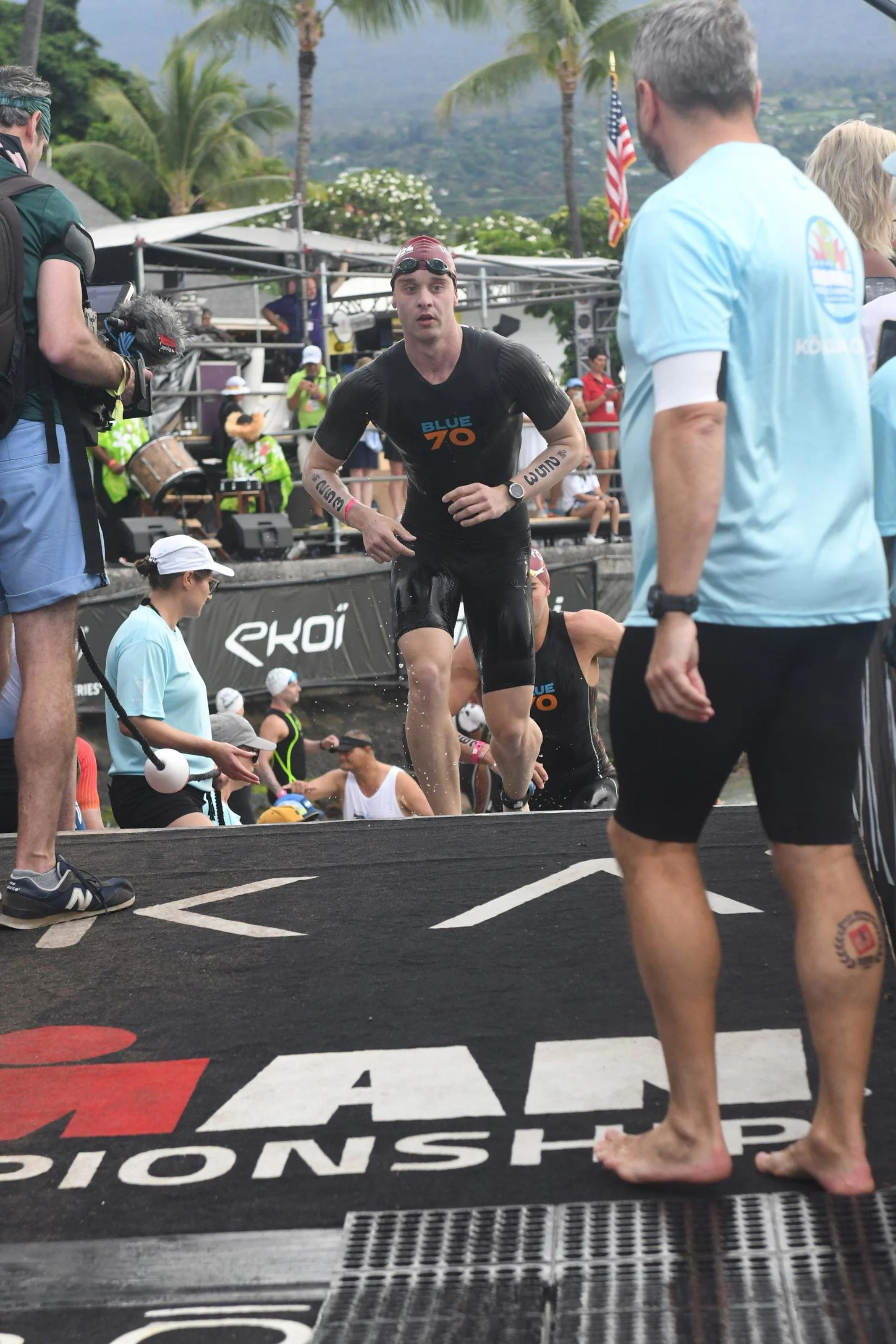 A male triathlete wearing a red swim cap, goggles, and a black wetsuit runs out of the water during a race, with spectators, officials, and tropical scenery in the background.