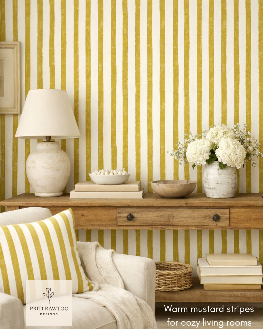 Interior of a cozy living room with a wooden console table against a mustard and white striped wallpaper. On the table are a white ceramic lamp, white books, a wooden bowl, a white vase with white hydrangeas, and a bowl with white decorative items. Part of a sofa with a striped mustard and white cushion and a cream throw blanket is visible in the foreground.