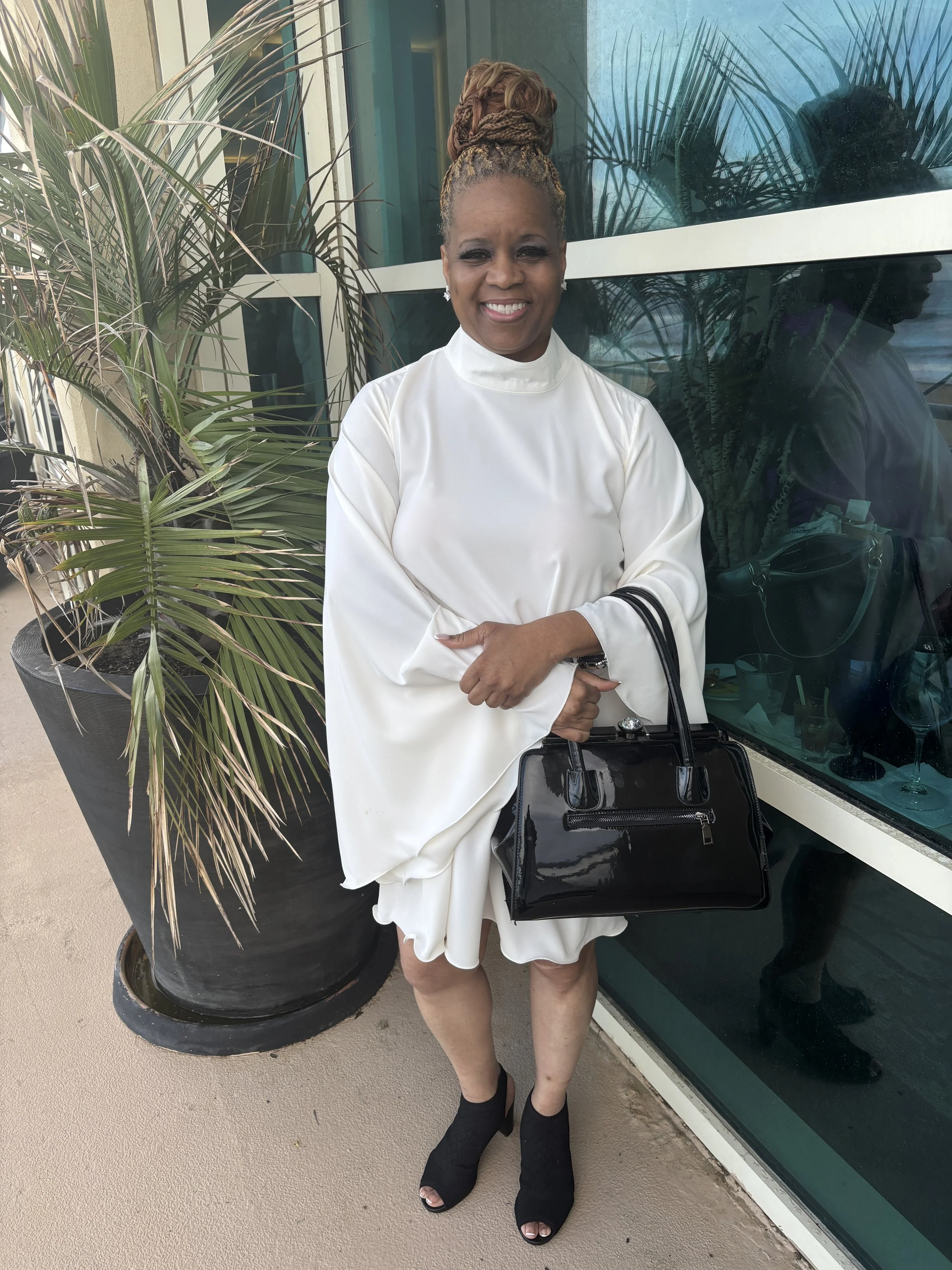 A woman with braided hair styled in a high bun is smiling and standing outdoors near a large potted plant and a glass window, holding a black handbag, wearing a white dress with black open-toe shoes.