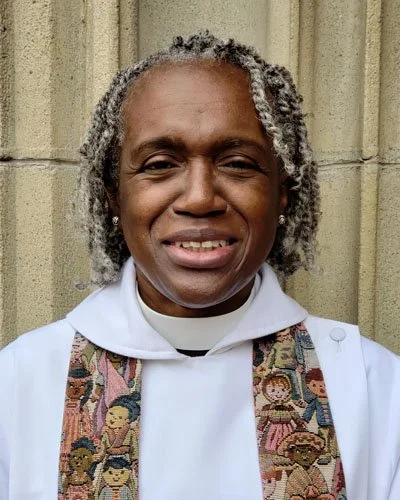 A woman dressed in religious clergy attire with grey, curly hair standing in front of a brick wall.
