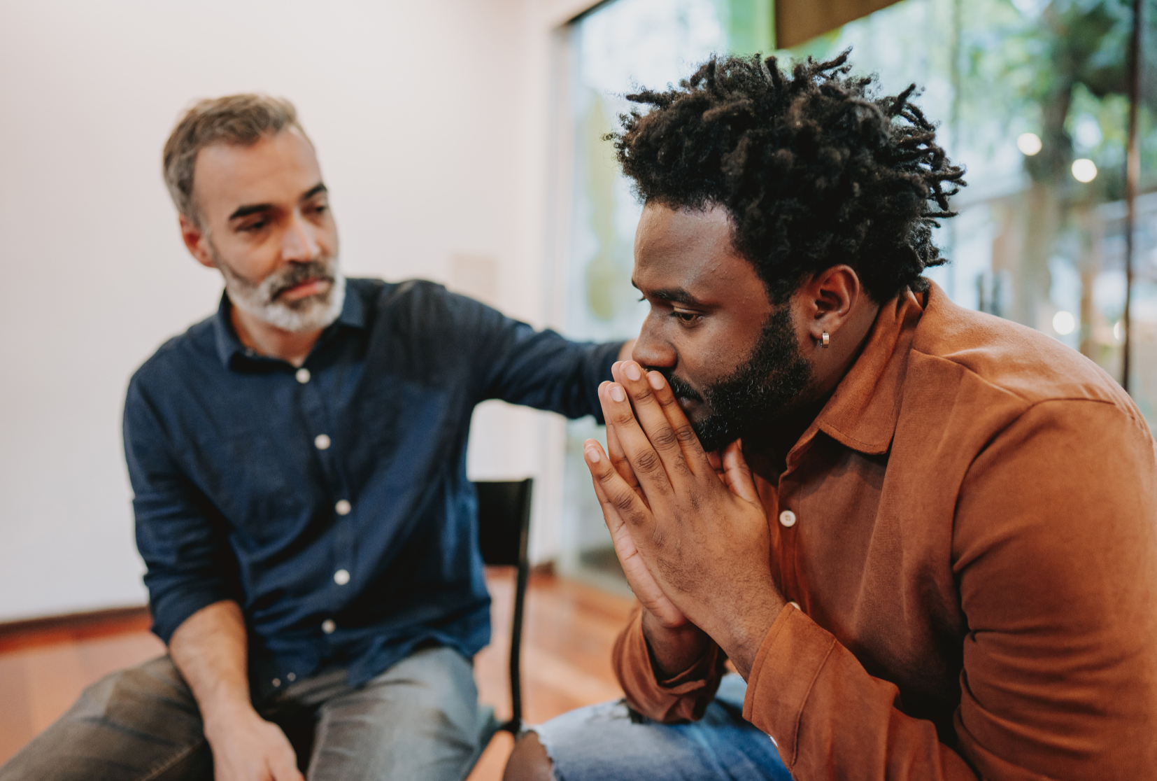 A man with a beard and gray hair comforting a younger man with curly hair, who appears to be upset or praying, in a room with a large window and greenery outside.