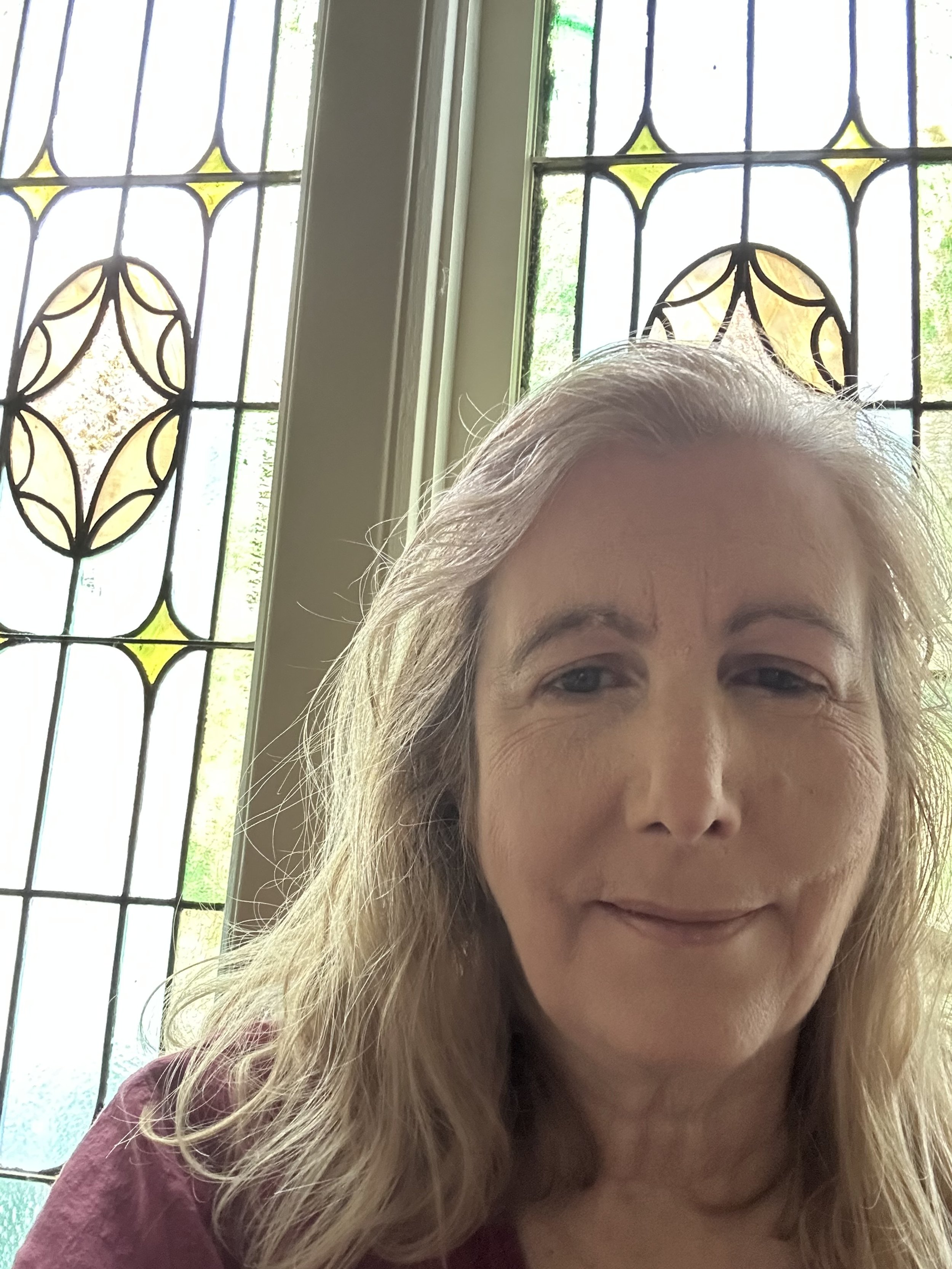A woman with light-colored hair taking a selfie indoors, with stained glass windows featuring geometric and floral designs in the background.
