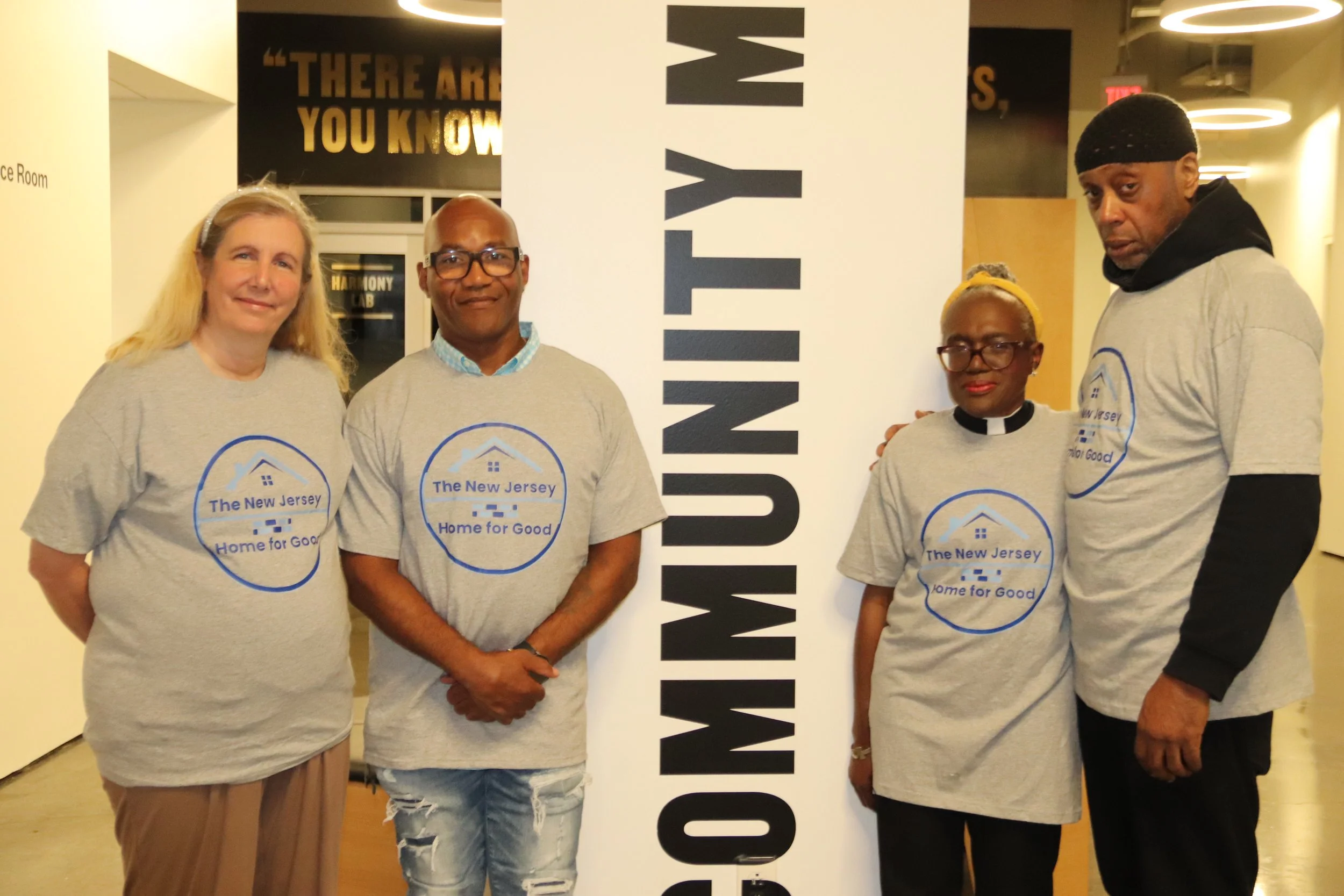 Four individuals standing indoors, each wearing a gray T-shirt with a logo for New Jersey Housing for Good. They are positioned next to a tall sign that says "COMMUNITY." The background includes a wall with a quote and a hallway with circular ceiling lights.