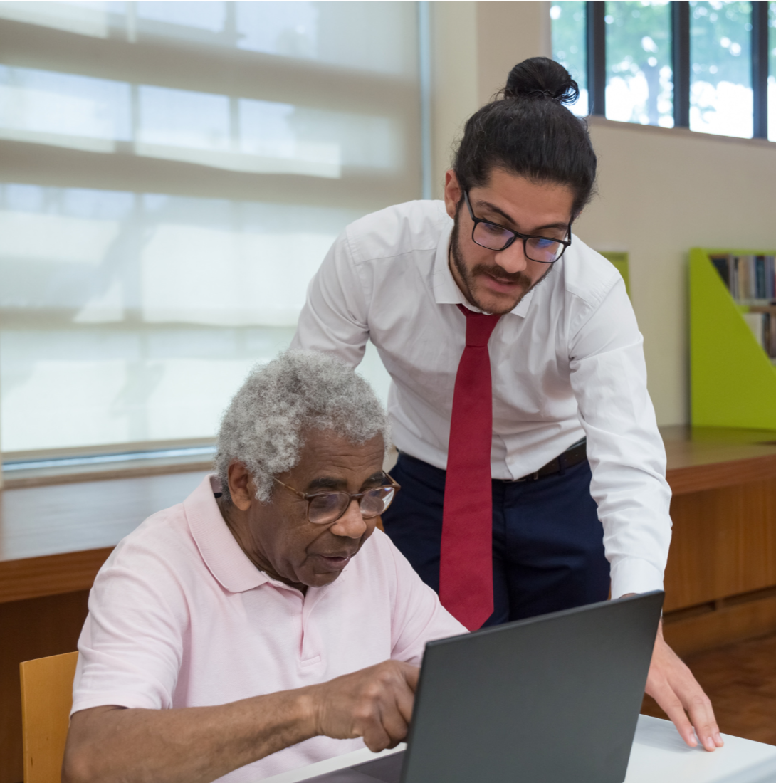 A young man with glasses and a man with gray curly hair and glasses viewed together looking at a laptop in a bright room with large windows and bookshelves.