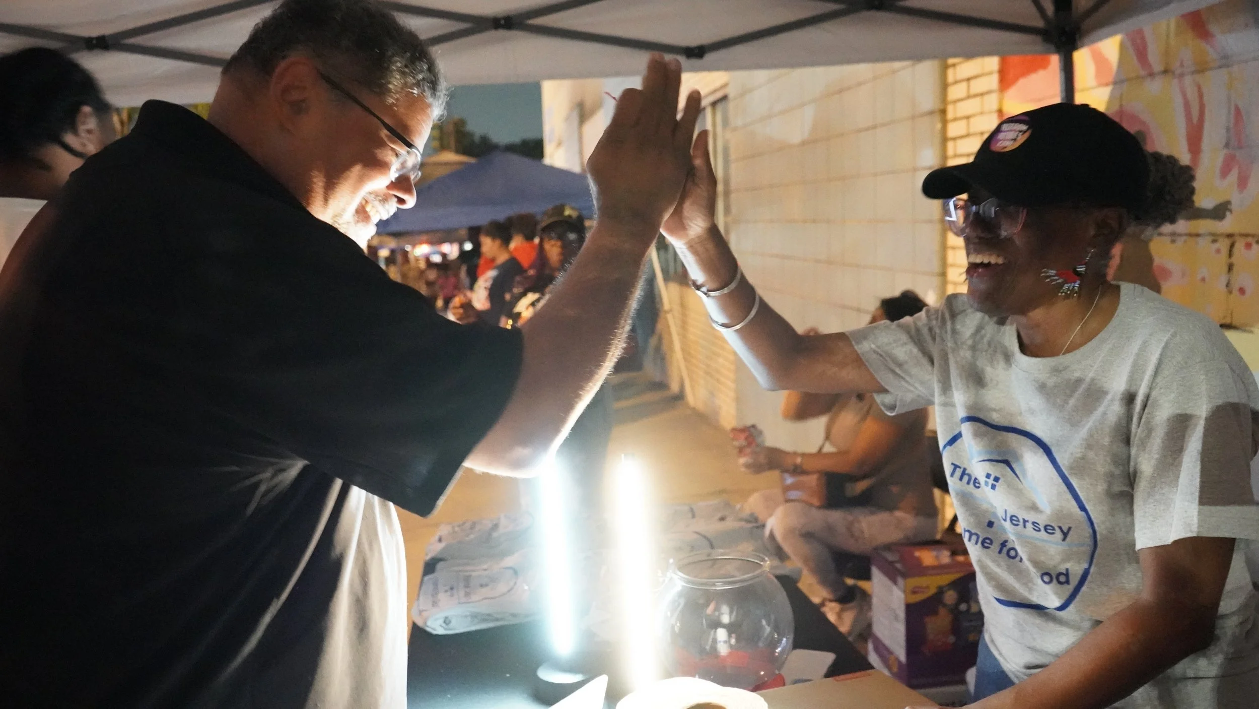A man and a woman high-five at an outdoor event, smiling. The man is wearing glasses and a black shirt, while the woman is wearing a gray shirt, glasses, and a cap. They are standing at a table with a fishbowl and some items, under a canopy with other people in the background.