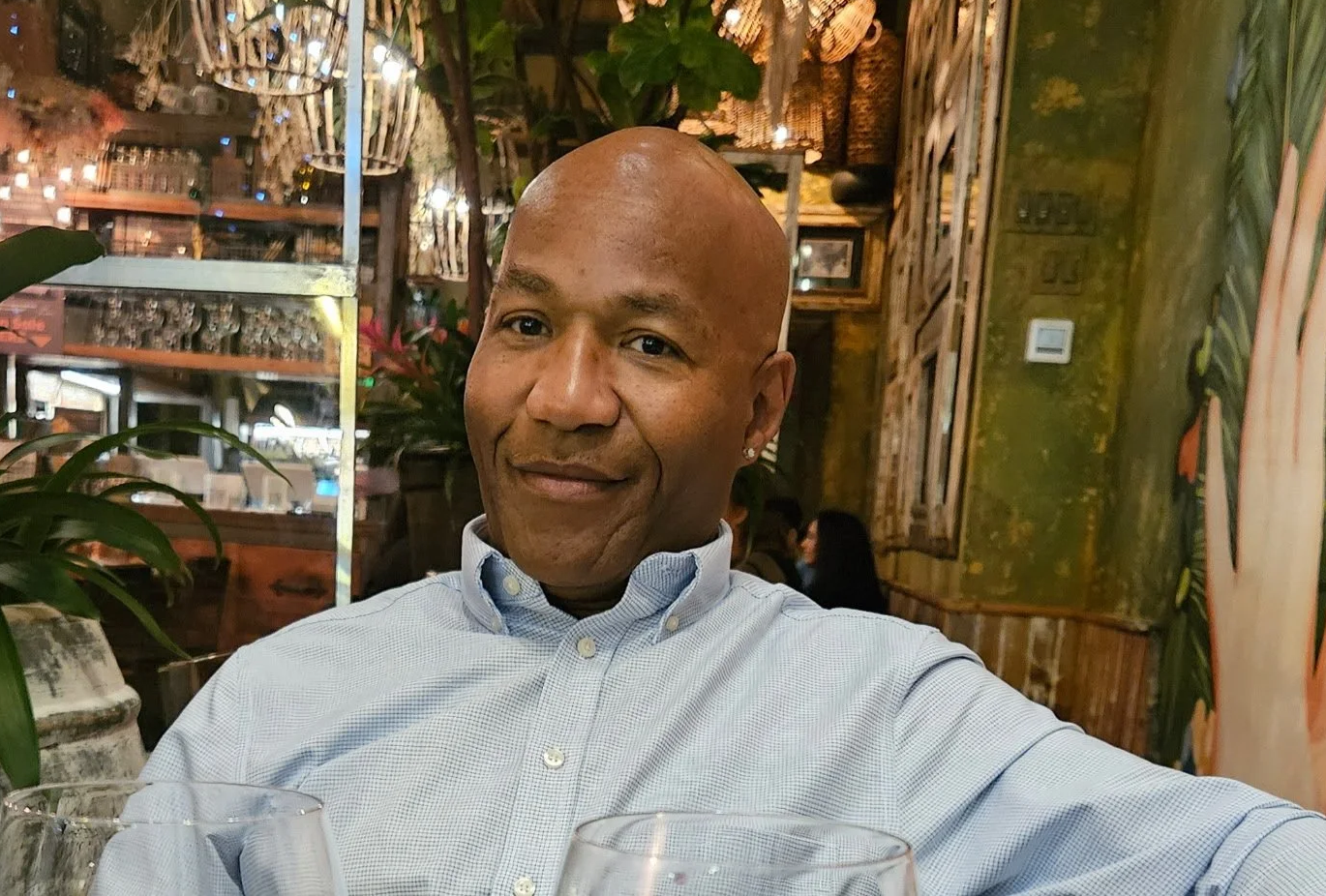 A man with a shaved head wearing a light blue button-down shirt sits in a restaurant with colorful, tropical-themed decor, including foliage, wooden furnishings, and warm lighting.
