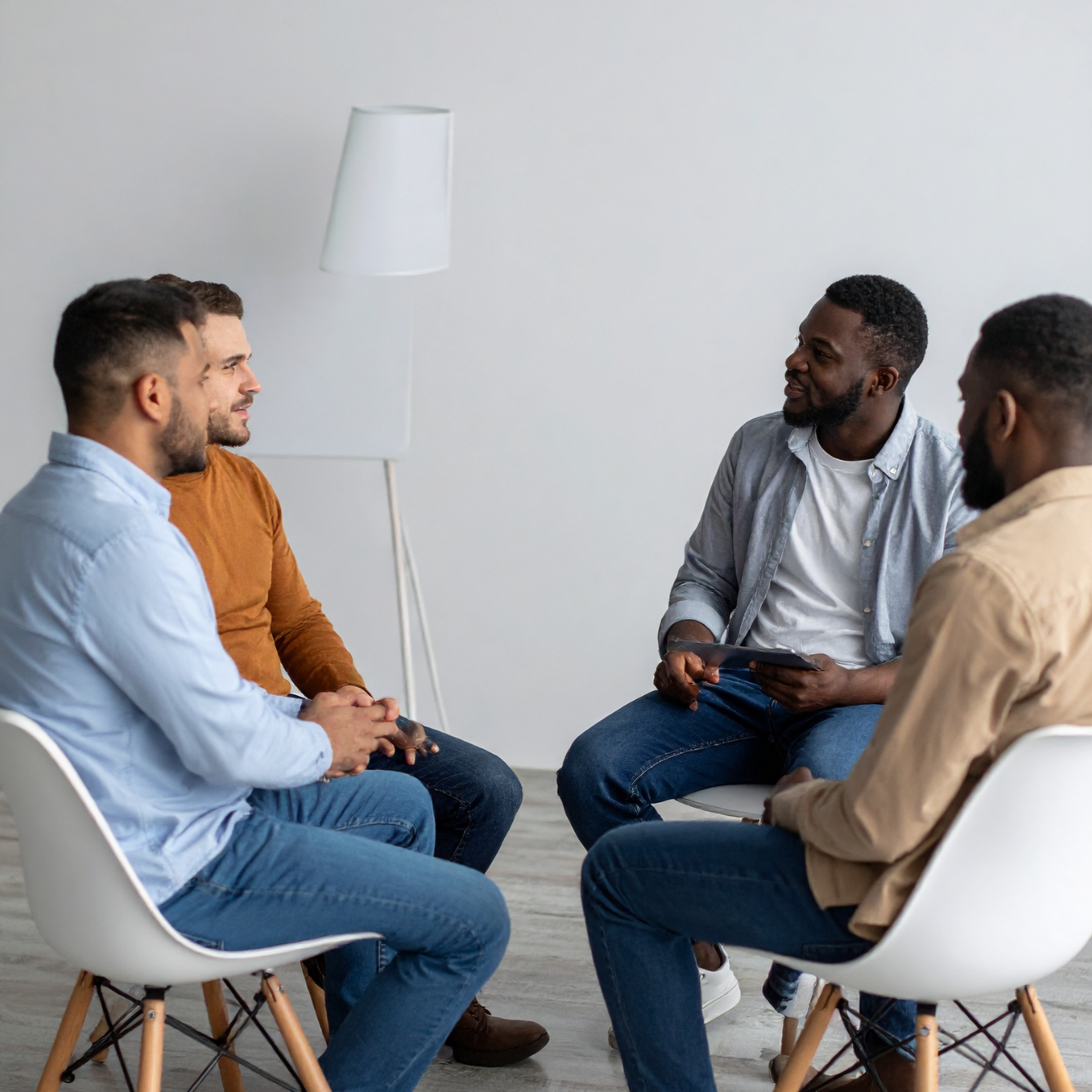 Five men sitting in a circle having a discussion in a bright, minimalist room with a white lamp in the background.