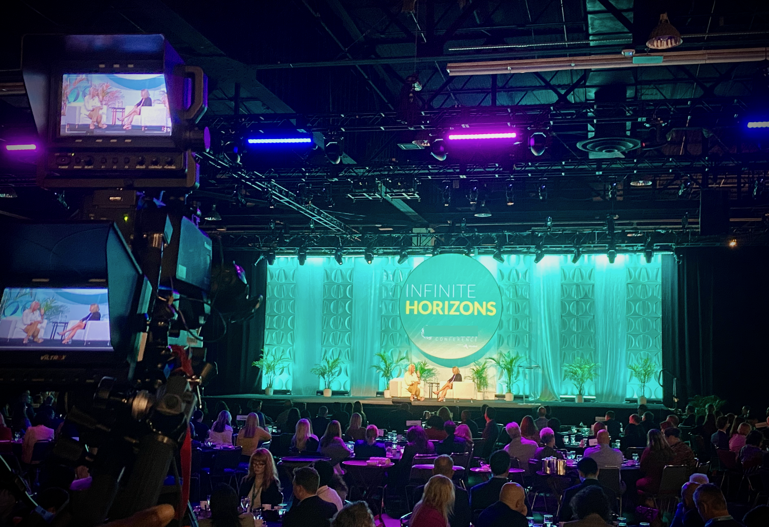 Large Scale Corporate General Session Event - Two people are seated on stage engaged in a conversation, framed by green plants and purple lighting overhead, with an audience seated in front.