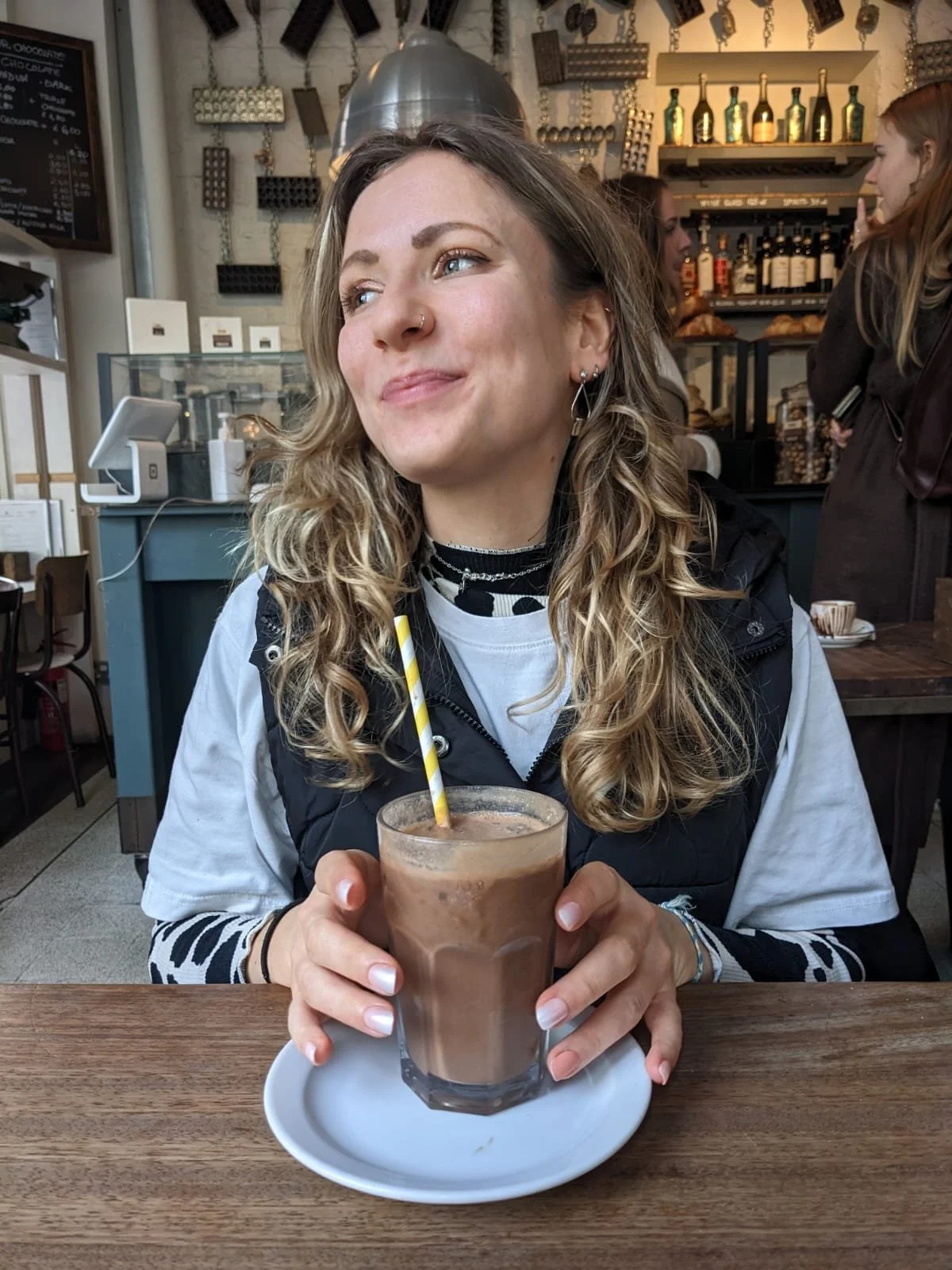 A woman with wavy blonde hair and earrings sitting at a wooden table, holding a glass of chocolate drink with a yellow-striped straw in a cafe.