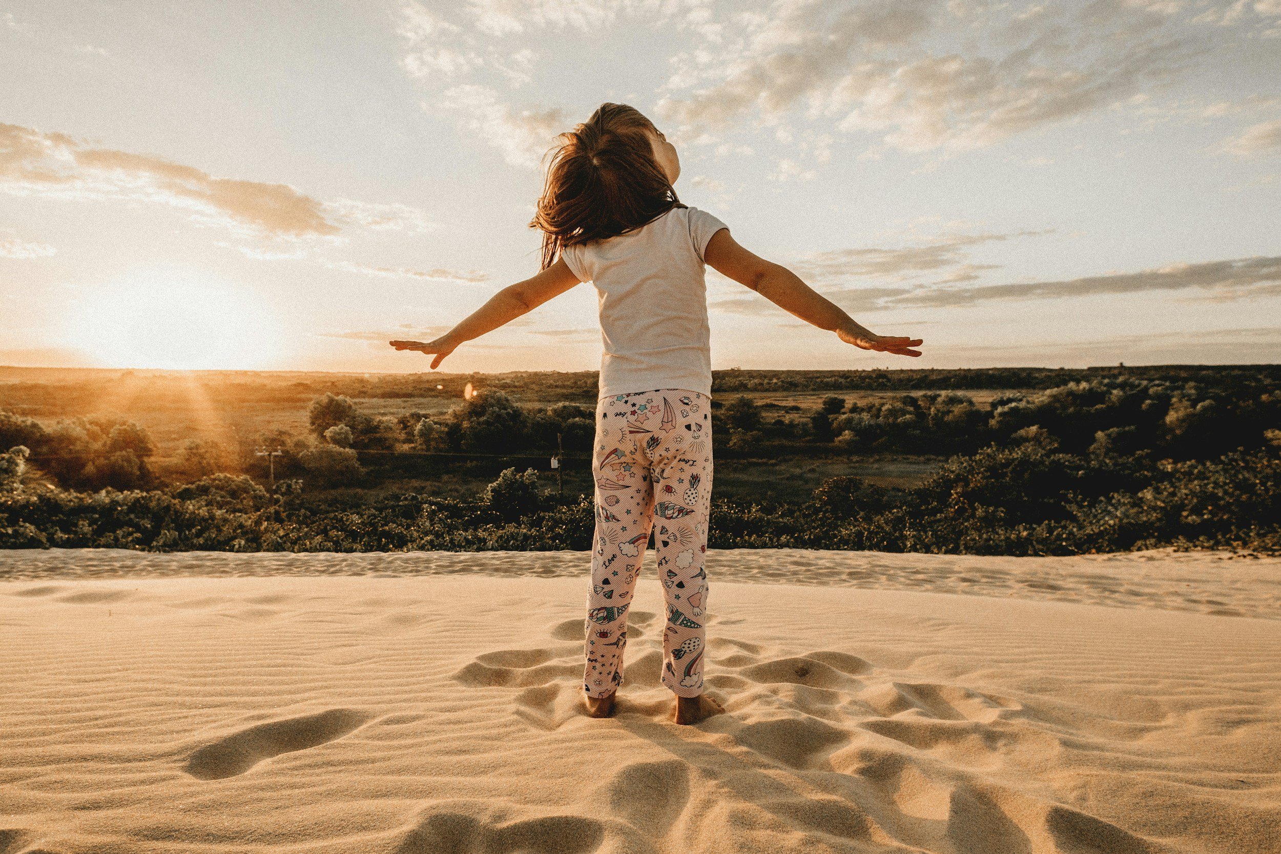 A girl with outstretched arms standing on sand dunes at sunset, facing away from the camera, with a landscape of trees and a partly cloudy sky in the background.