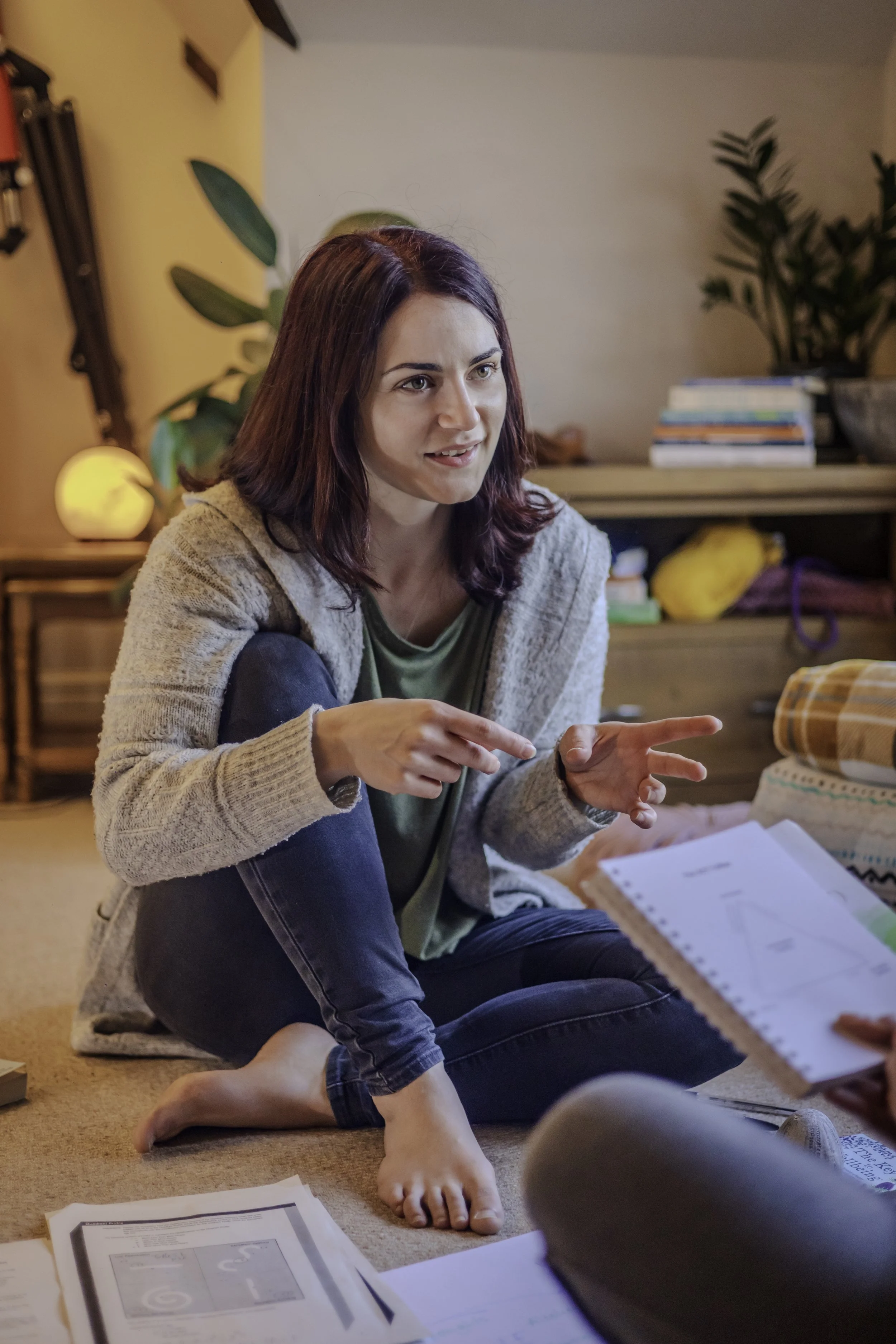A woman with shoulder-length dark hair having a conversation indoors, sitting on the carpeted floor surrounded by papers and notebooks, with a cozy background including a wooden cabinet, a plant, and a glowing lamp.