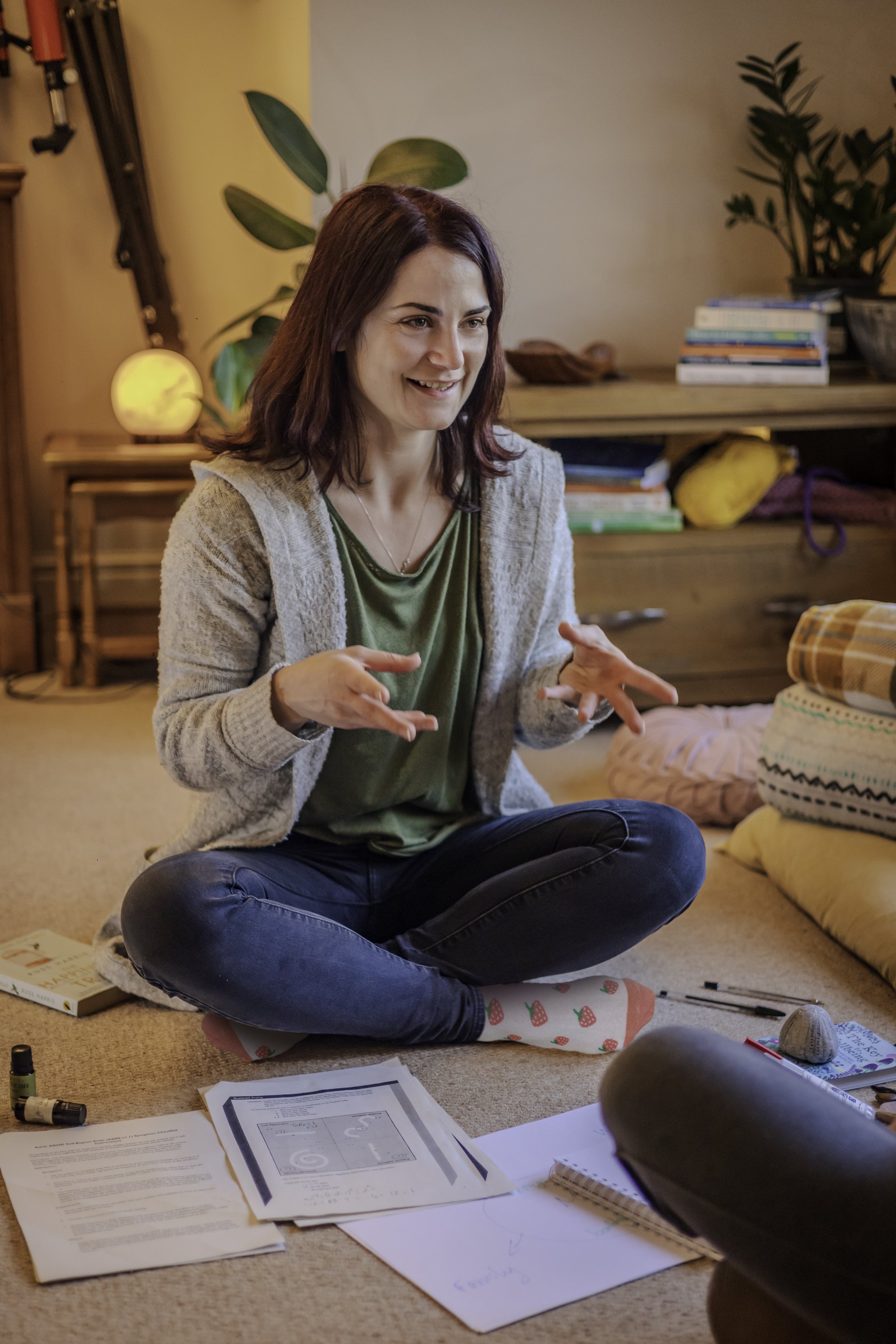 Woman sitting cross-legged on carpet, gesturing with hands, smiling, surrounded by papers and objects, in a cozy room with plants and shelves.