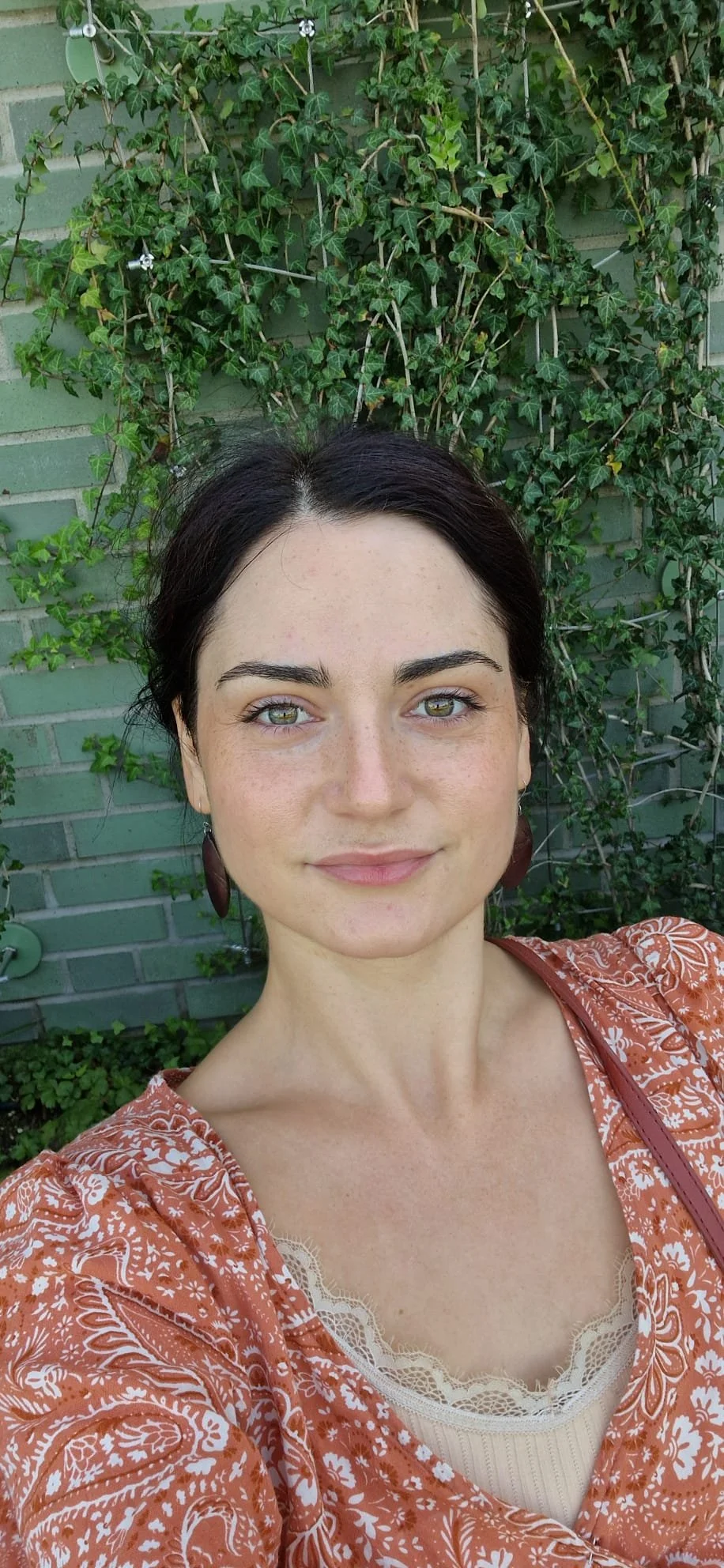 A woman taking a selfie outdoors, standing in front of a green brick wall covered with lush green ivy.
