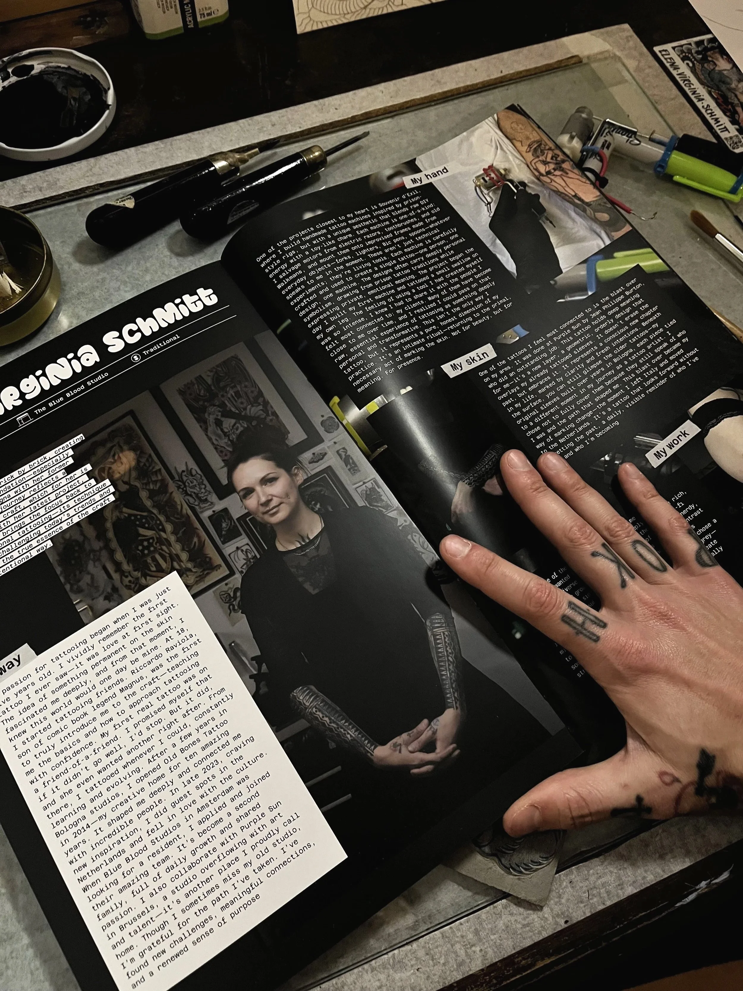 Open magazine on a tattoo artist's workspace, showing an article about a woman with tattoos, surrounded by tattooing tools, ink, and artwork.