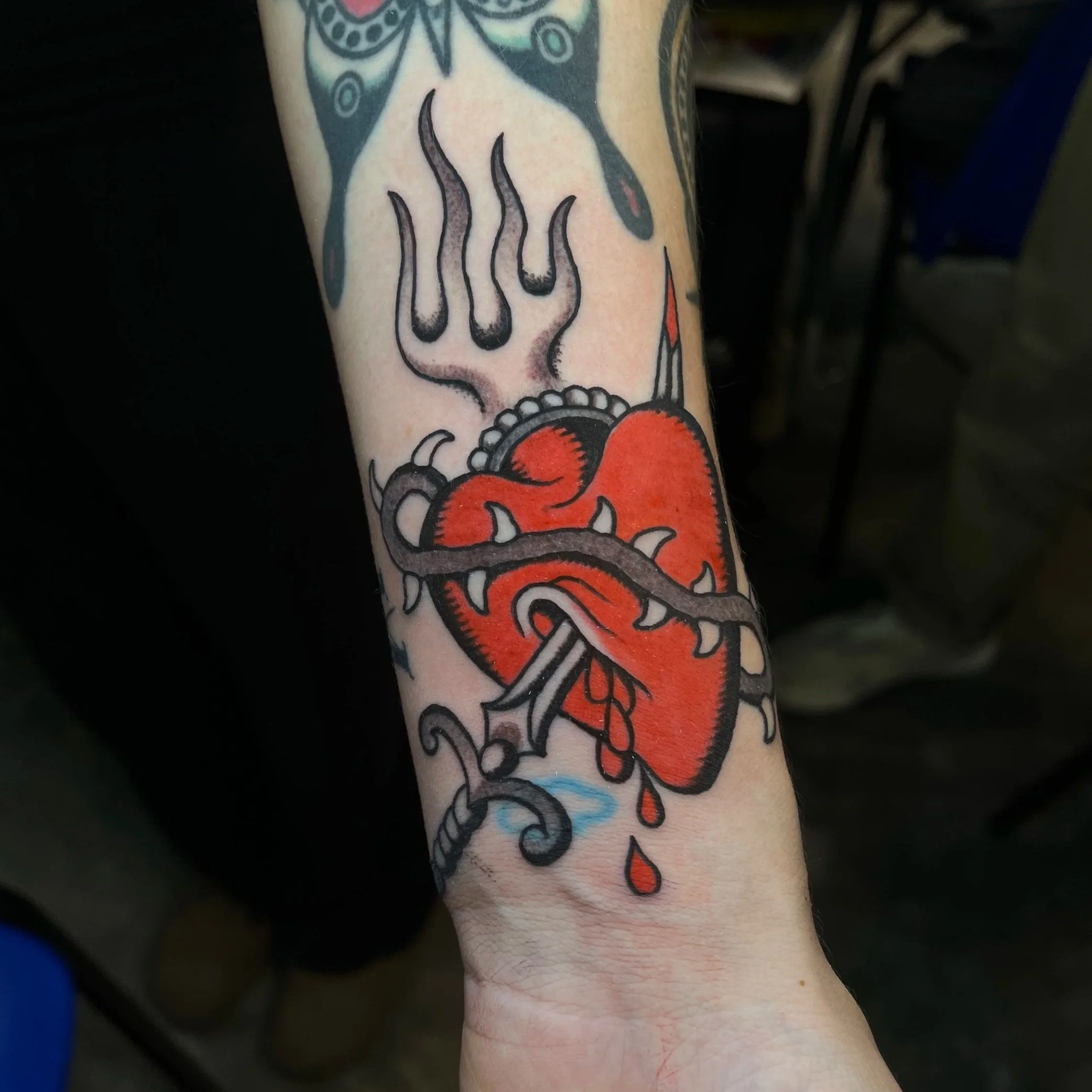 Tattoo of a red heart pierced by a dagger with a thorny vine wrapping around it, black flames above the heart, and red blood drops below.