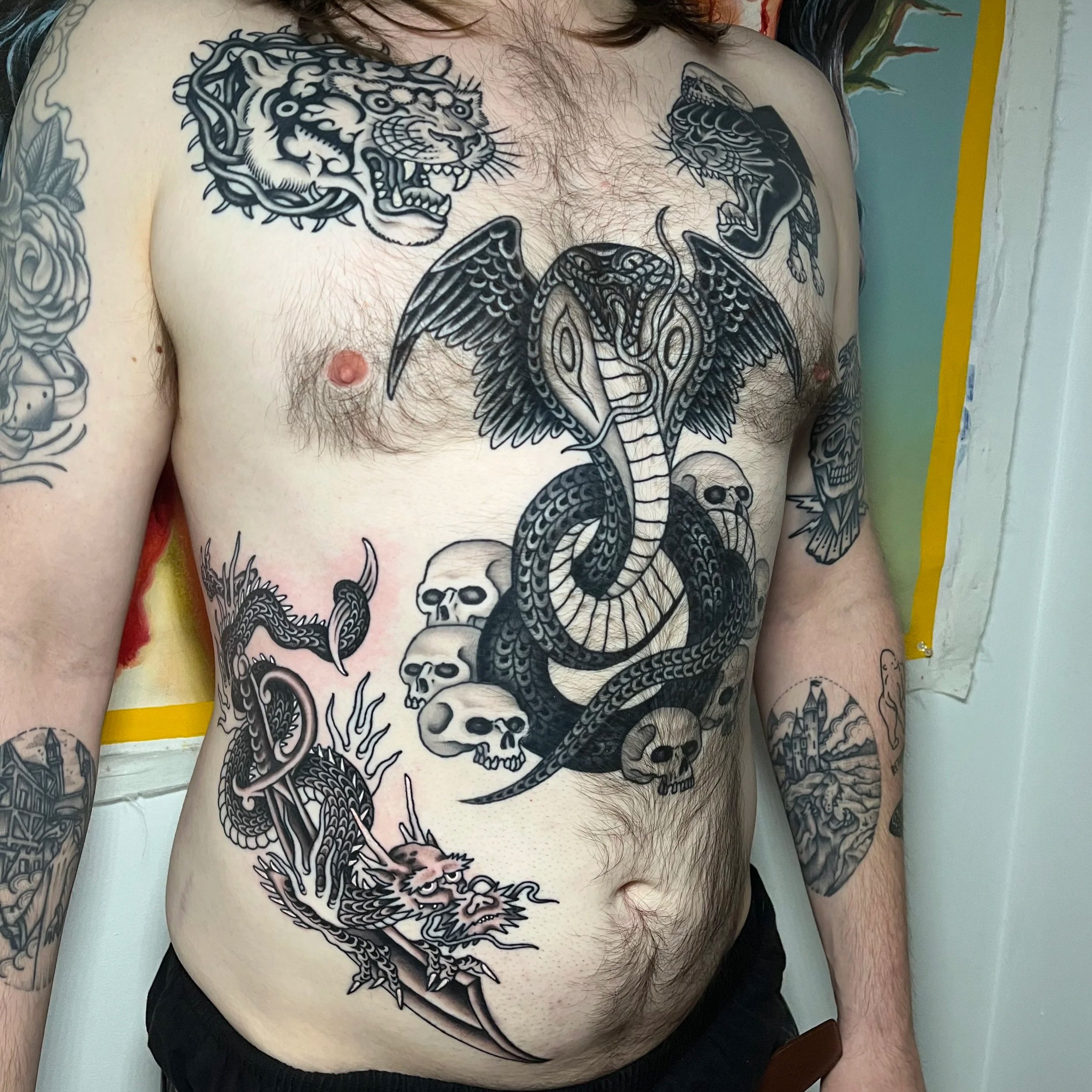A man's chest tattoo featuring a large snake with wings, surrounded by skulls and smaller dragons, with additional tattoos visible on his arms.