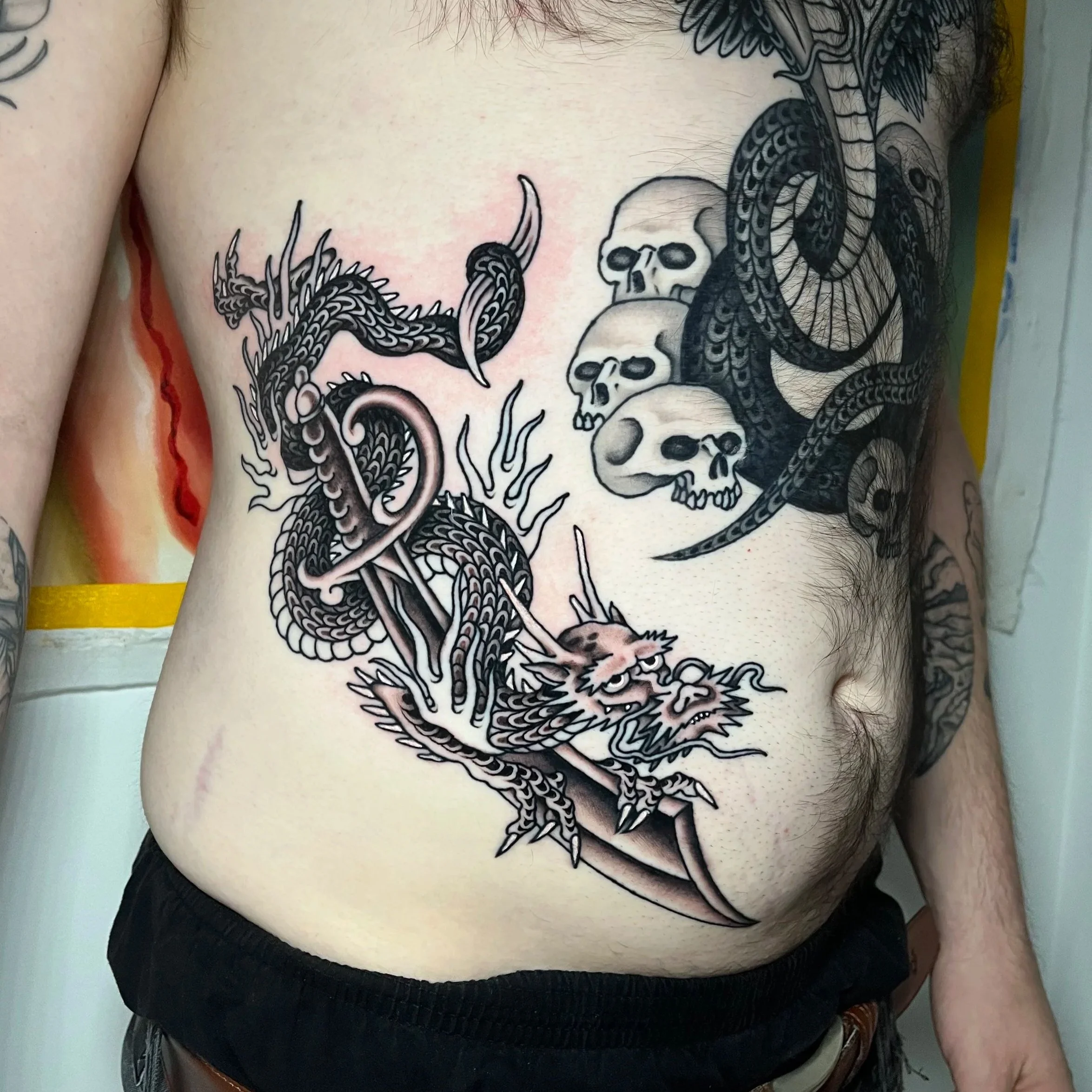 A tattoo of a dragon on the person's stomach, with a snake's head and multiple skulls nearby.