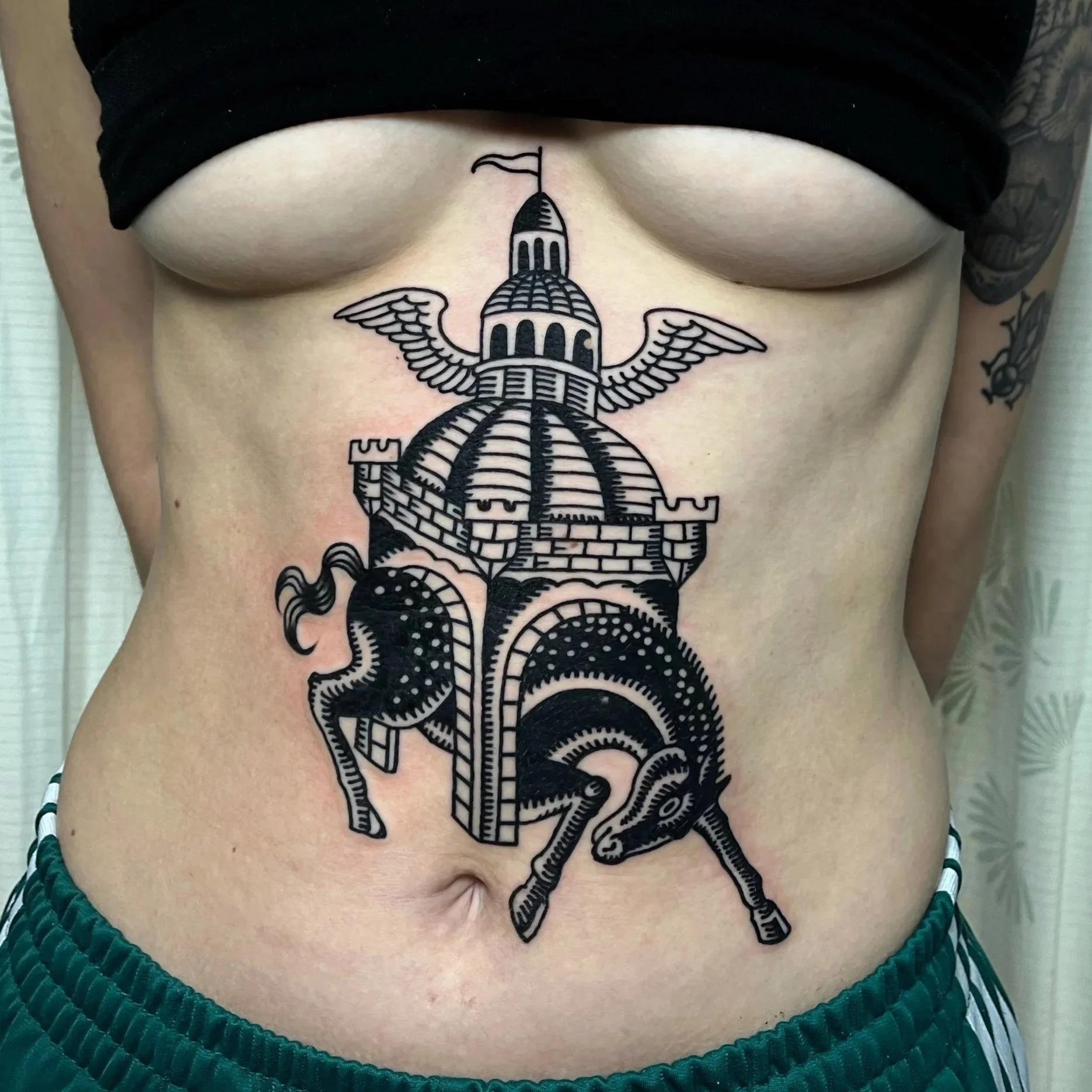 Black tattoo on person's abdomen of a horse with a castle and wings.