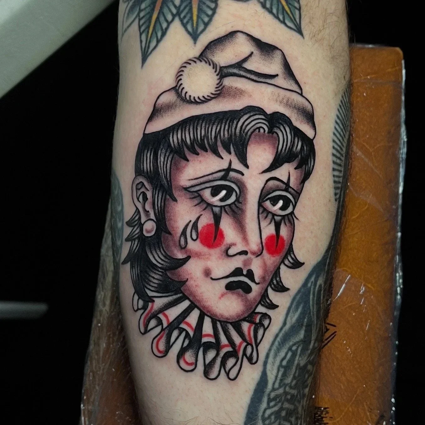 Tattoo of a sad clown with tears, red cheeks, dark hair, wearing a collar with red accents.