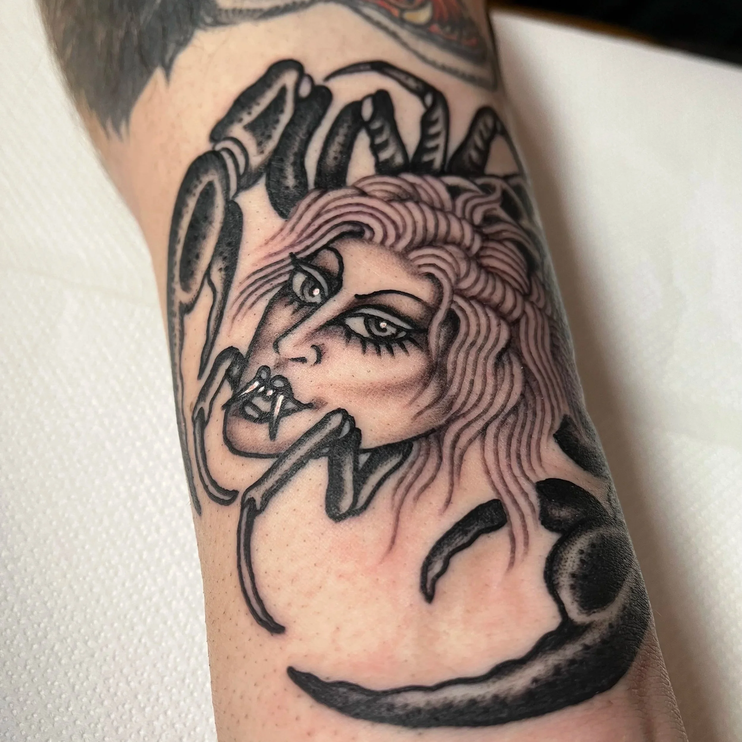 Tattoo of a woman's face with long wavy hair and large eyes, surrounded by spider legs, on a person's arm.