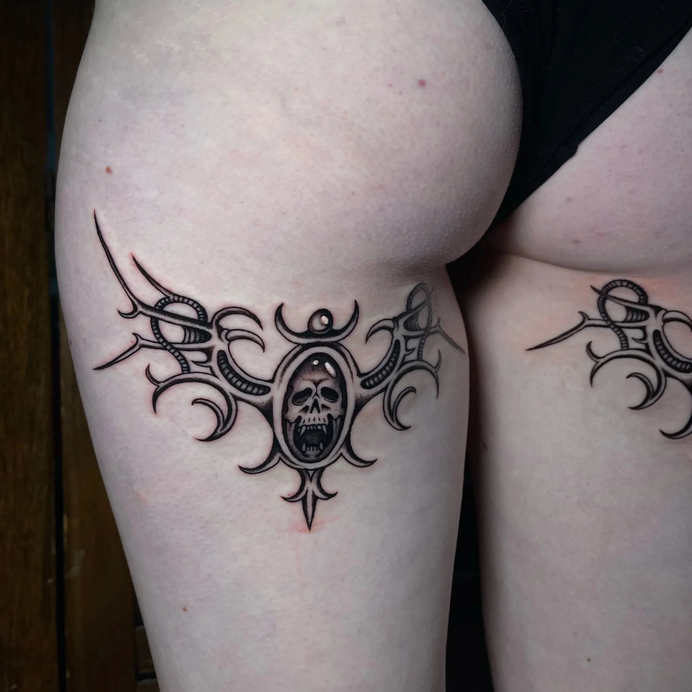 Tattoo of a skull with an oval around it, surrounded by curved and pointed lines, on upper thigh near the hip.