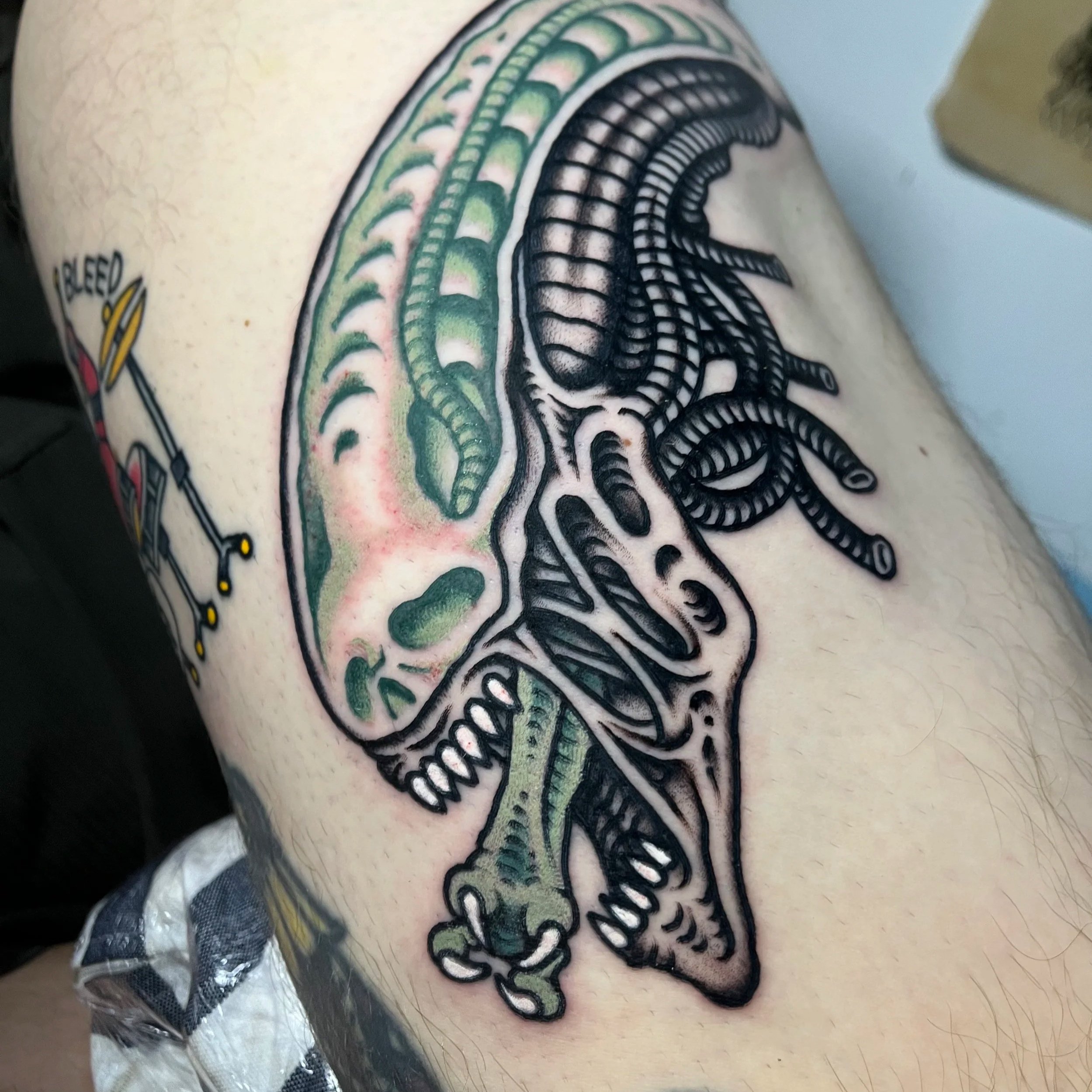 Colorful tattoo of alien skull with green and black details on a person's upper arm.