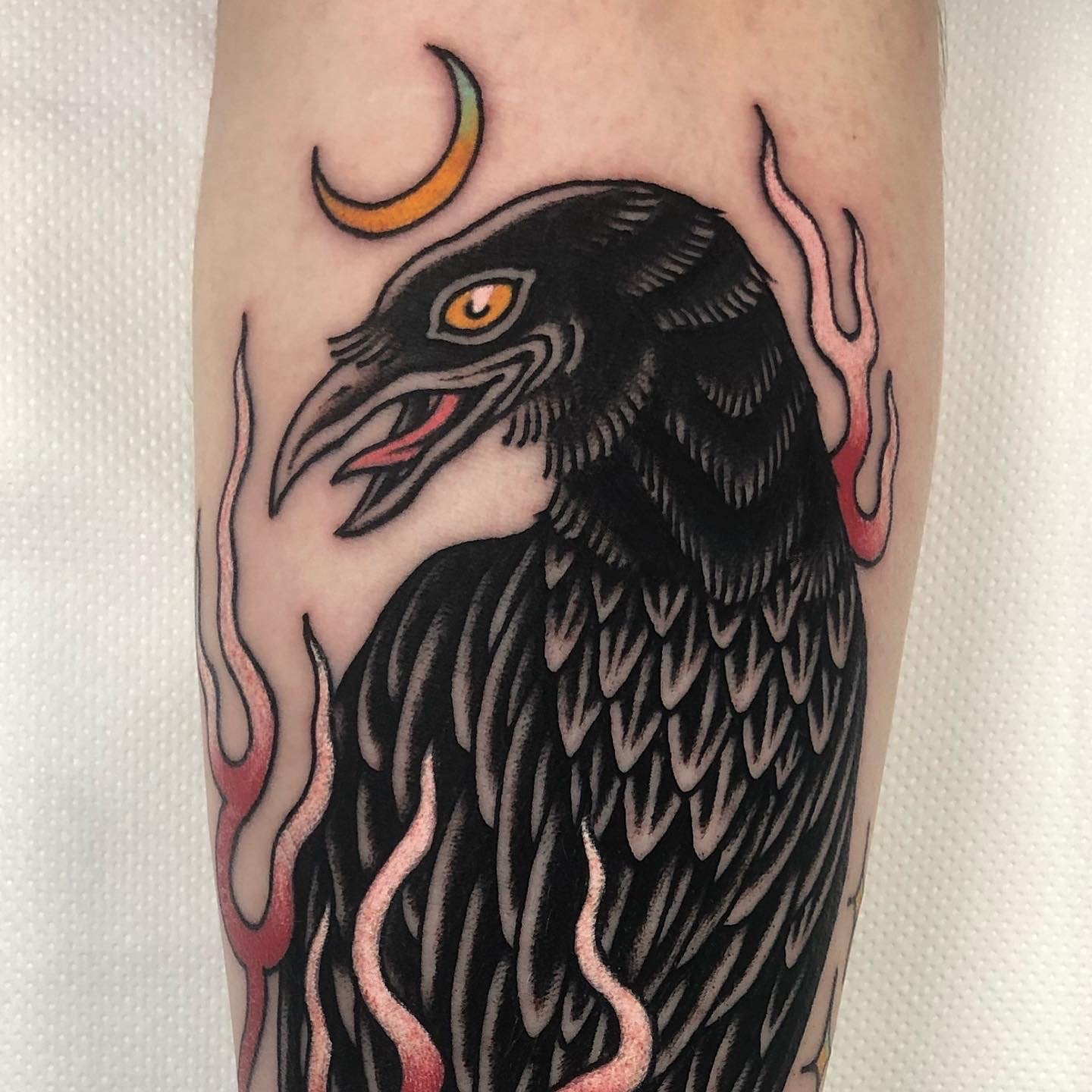 Tattoo of a black crow with orange eyes, surrounded by pink and red flames, with a small yellow crescent moon above it.