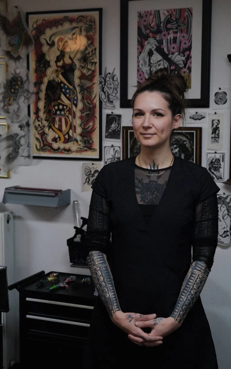 A woman with tattoos on her arms, chest, and face standing in front of tattoo artwork on a wall, wearing a black top with sheer sleeves and a necklace.