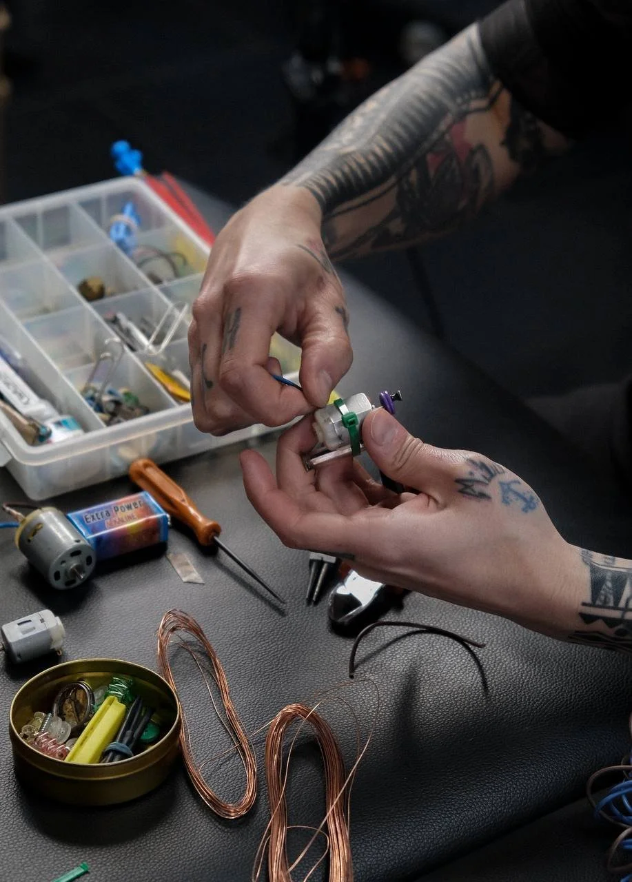 Person with tattoos assembling a jail tattoo machine , surrounded by tools and parts.