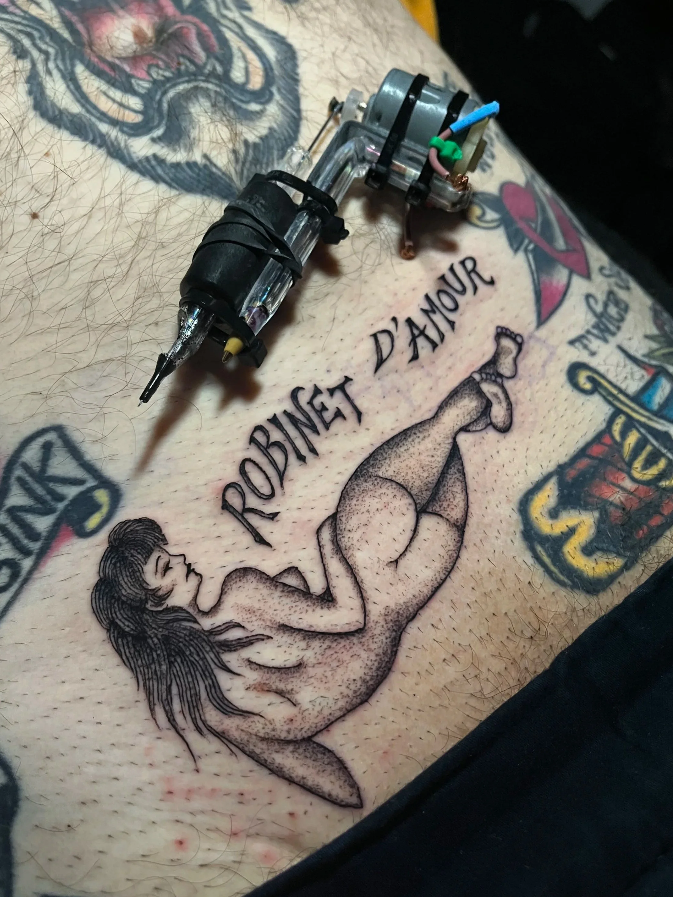 A tattoo of a nude woman with long hair, lying on her side, with the phrase "ROBINET D'AMOUR" above her. The tattoo is on a person's belly with additional colorful tattoos visible nearby.