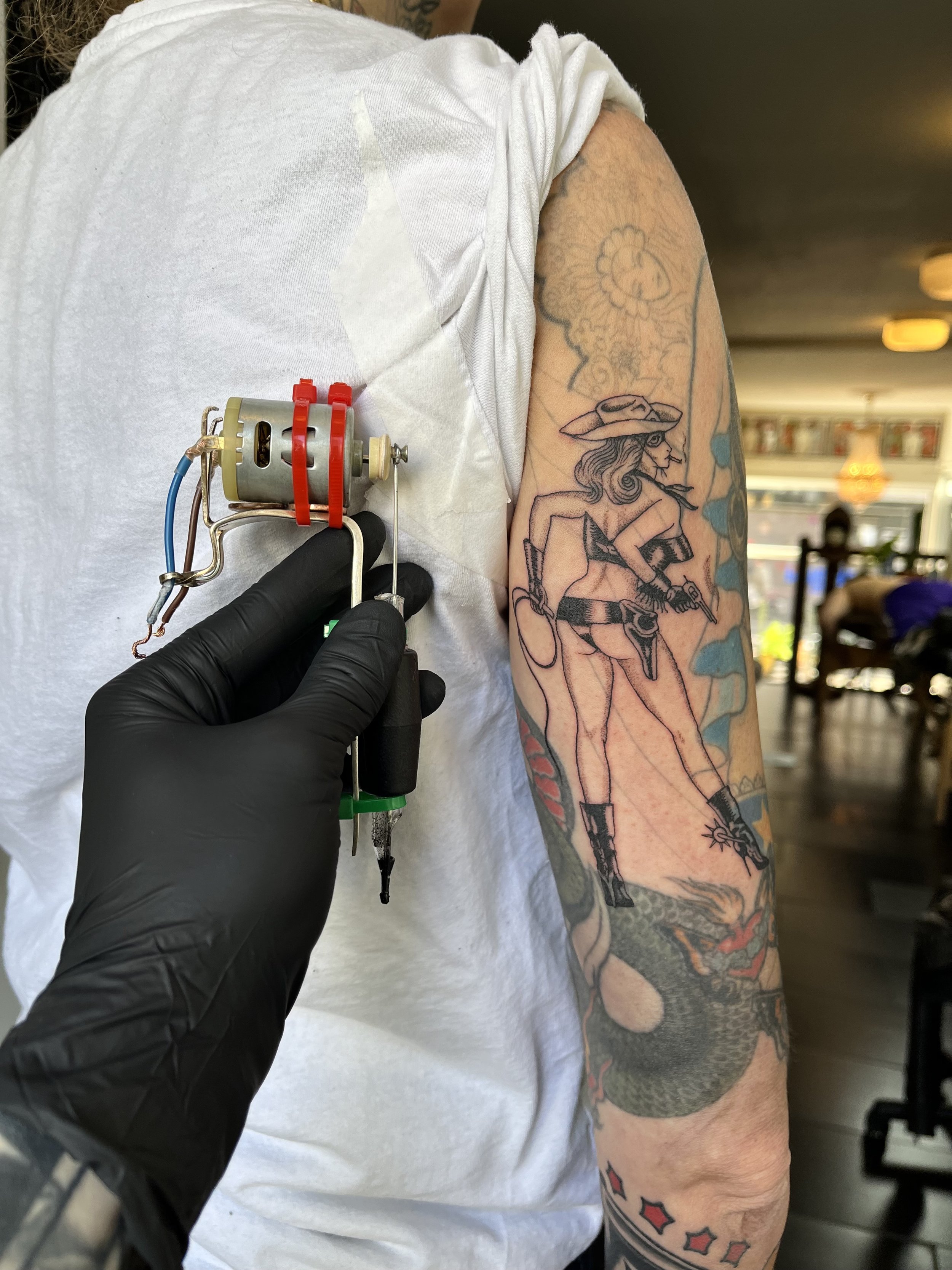 A person getting a tattoo of a cowgirl holding a gun on their upper arm, with a dragon tattoo below. The tattoo artist, wearing black gloves, is working on the tattoo using a jail tattoo machine