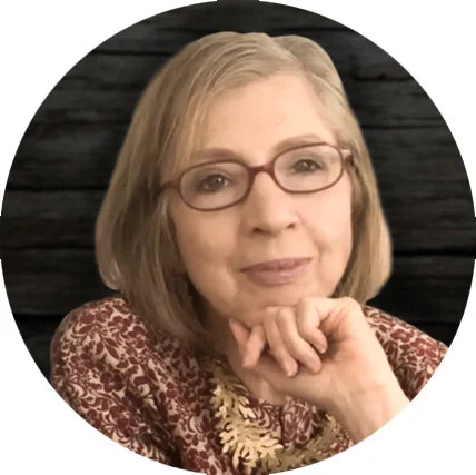 A woman with blonde hair and glasses, wearing a patterned blouse, resting her chin on her hand against a wooden background.
