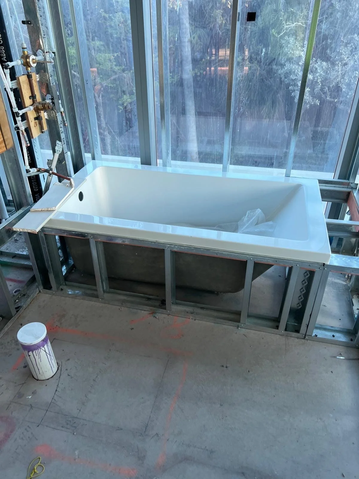 A bathroom under construction with a white bathtub inside a metal frame, surrounded by glass windows. There is a small wooden platform with plumbing fixtures on the left side of the bathtub.