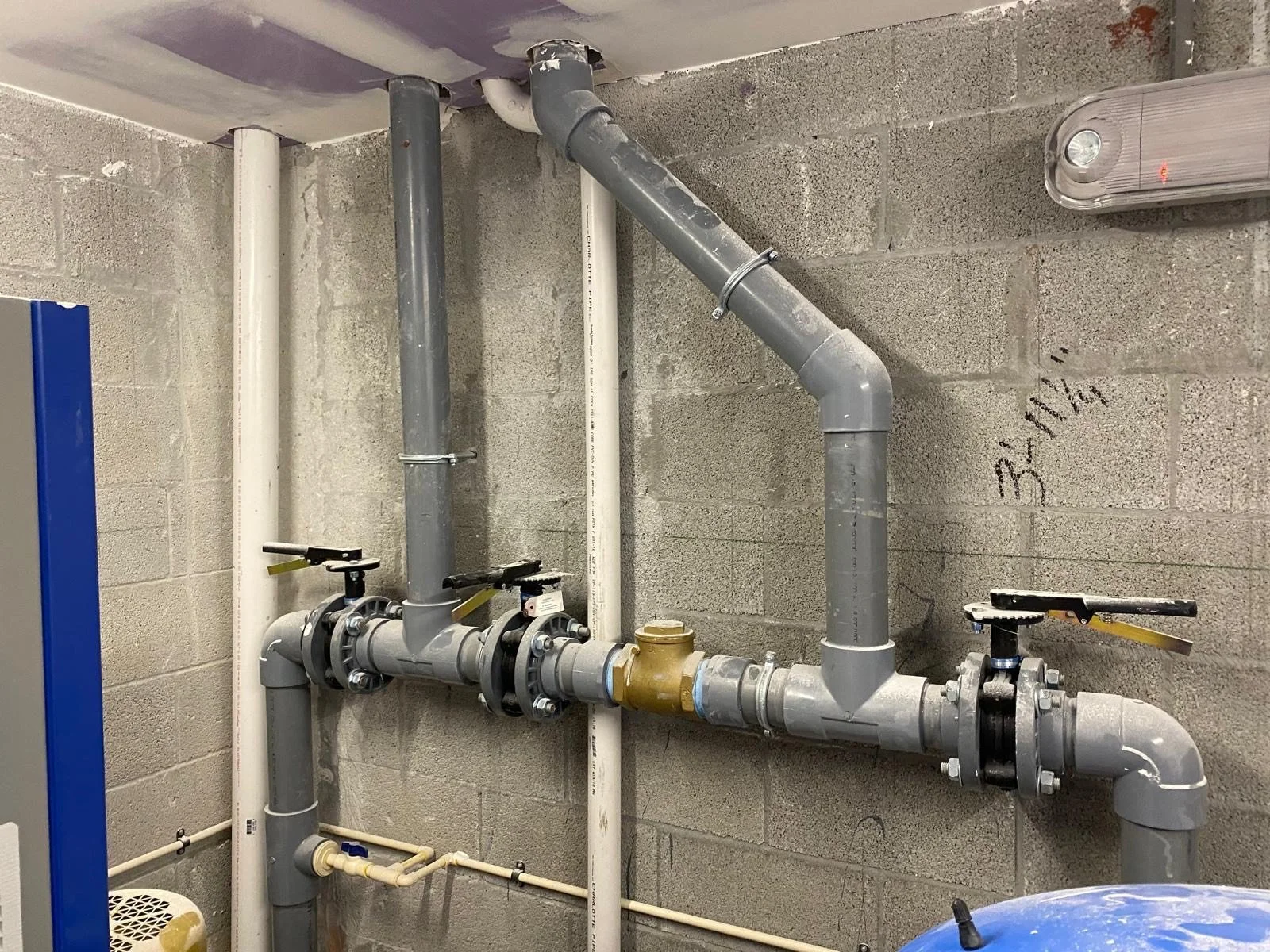 A plumbing wall with gray pipes, white pipes, and a bronze valve, with two black handles and two yellow tags, installed on a concrete wall with markings.