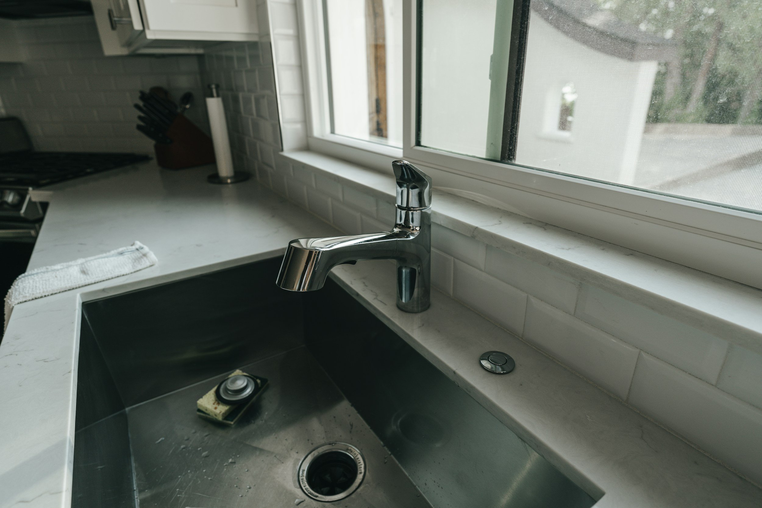 Stainless steel kitchen sink with faucet, soap dish, and dish sponge, beside a window with a white sill and brick backsplash.