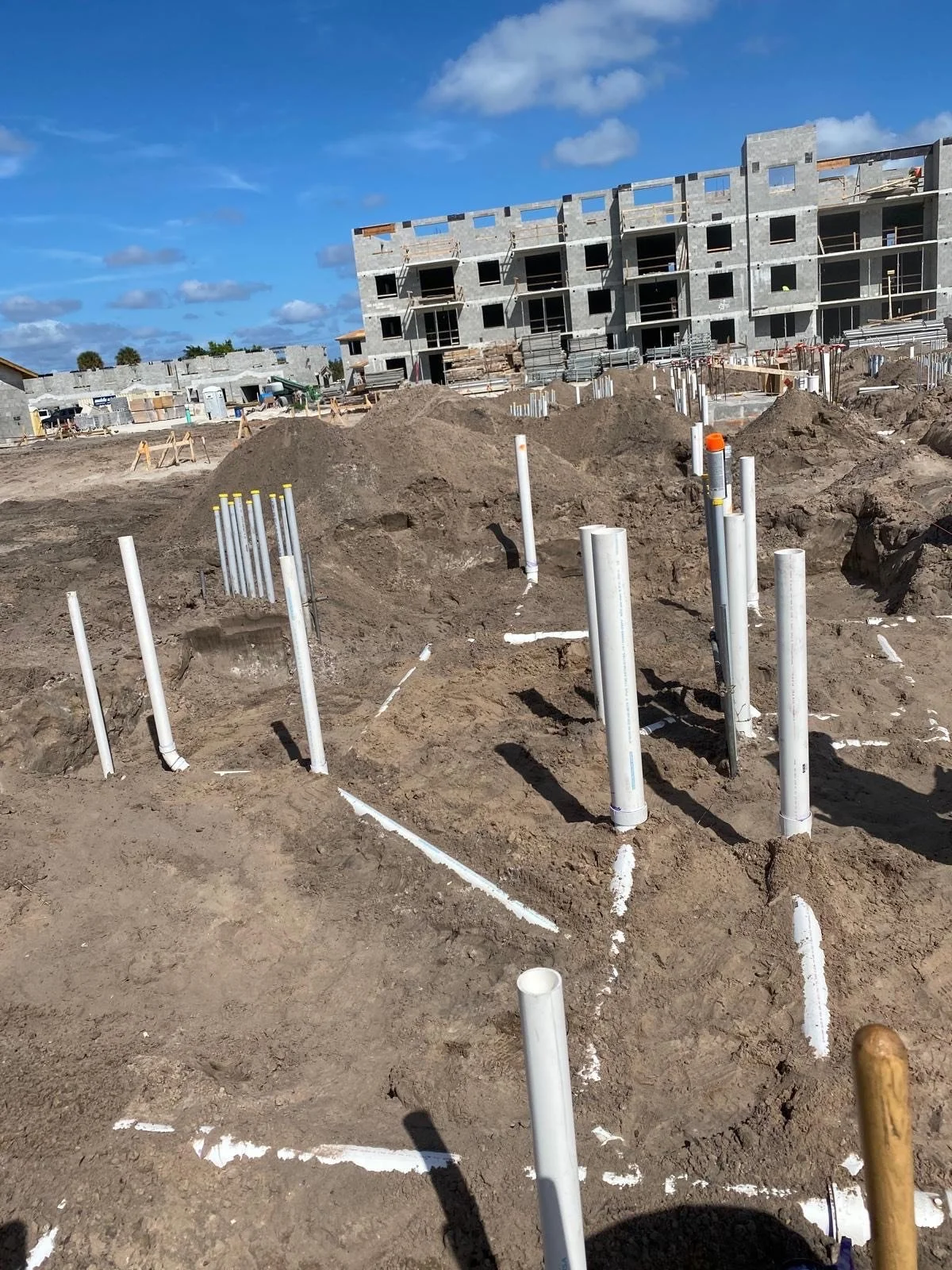 Construction site with exposed pipe foundations and building under construction in the background under a partly cloudy sky.
