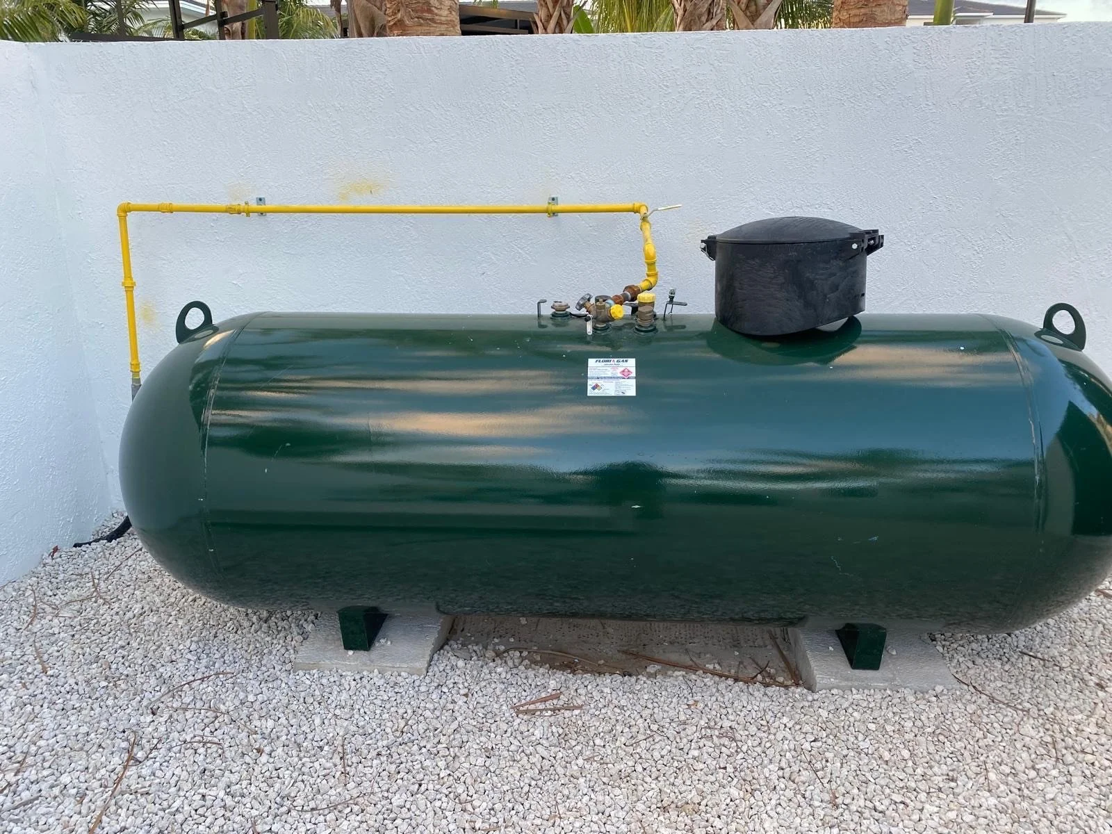 Green horizontal propane tank with yellow gas line, black regulator, and black cover on top, placed on gravel ground against a white wall.