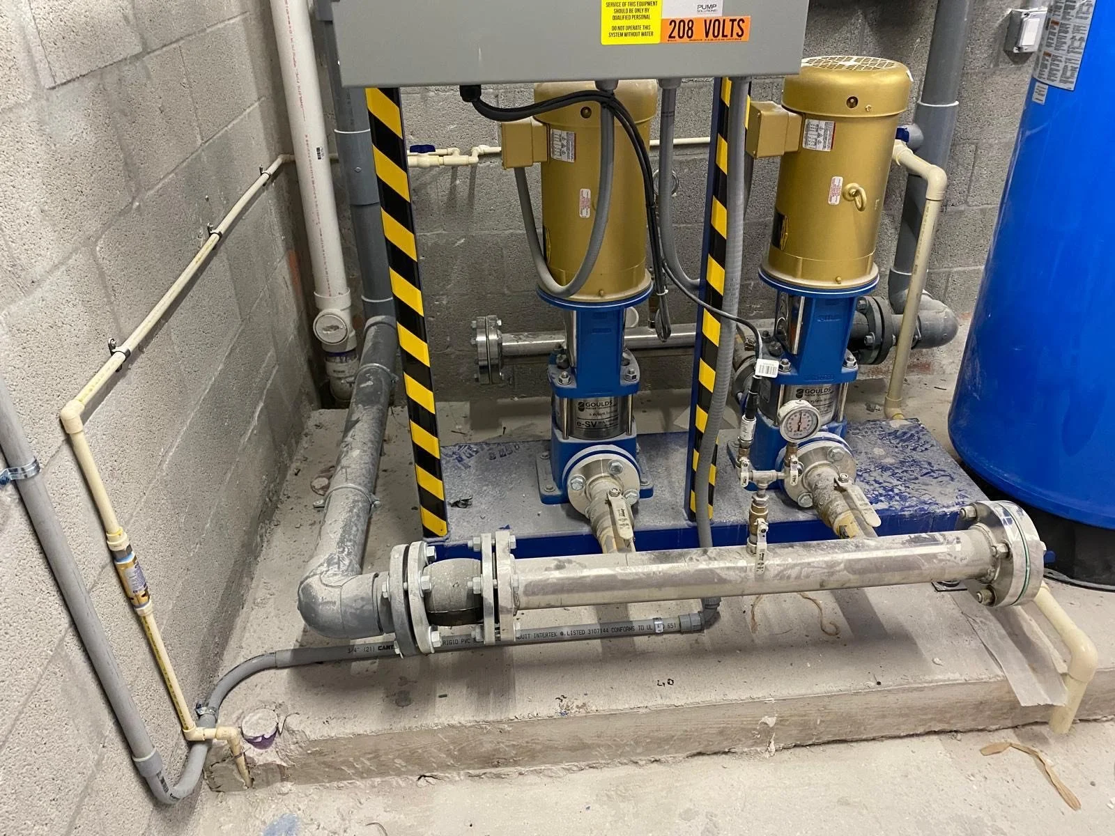 Industrial plumbing system with pipes, valves, gauges, and two yellow and blue pumps on a concrete floor.